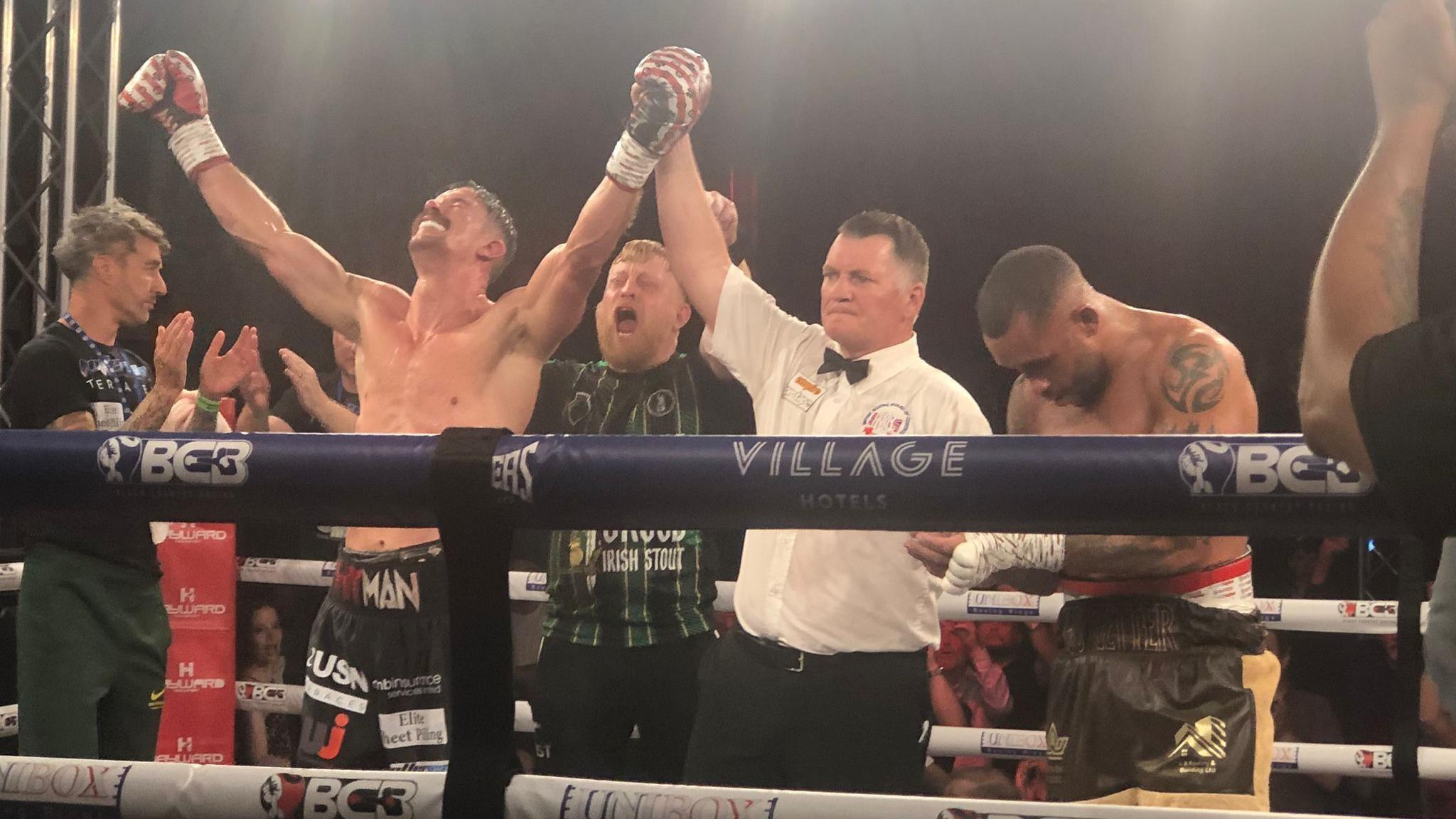 Two boxers stand in a boxing ring with a referee in white shirt and black bow tie between them. The boxer on the left looks up and holds his hands aloft as he is declared the winner.