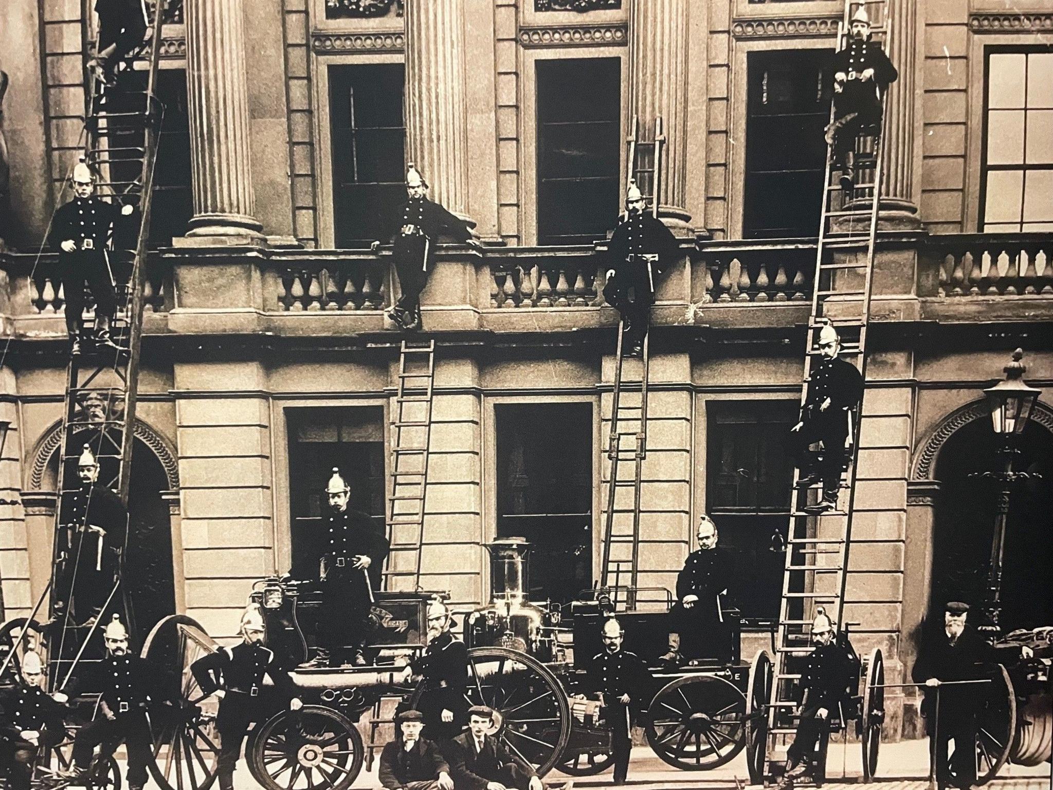 An archive photo of Inverness fire brigade shows about a dozen firefighters posing for the camera in from of a large stone building. The firefighters are standing on or around an old fire appliance. Four of the firefighters are posing leaning half way up ladders propped up against the building