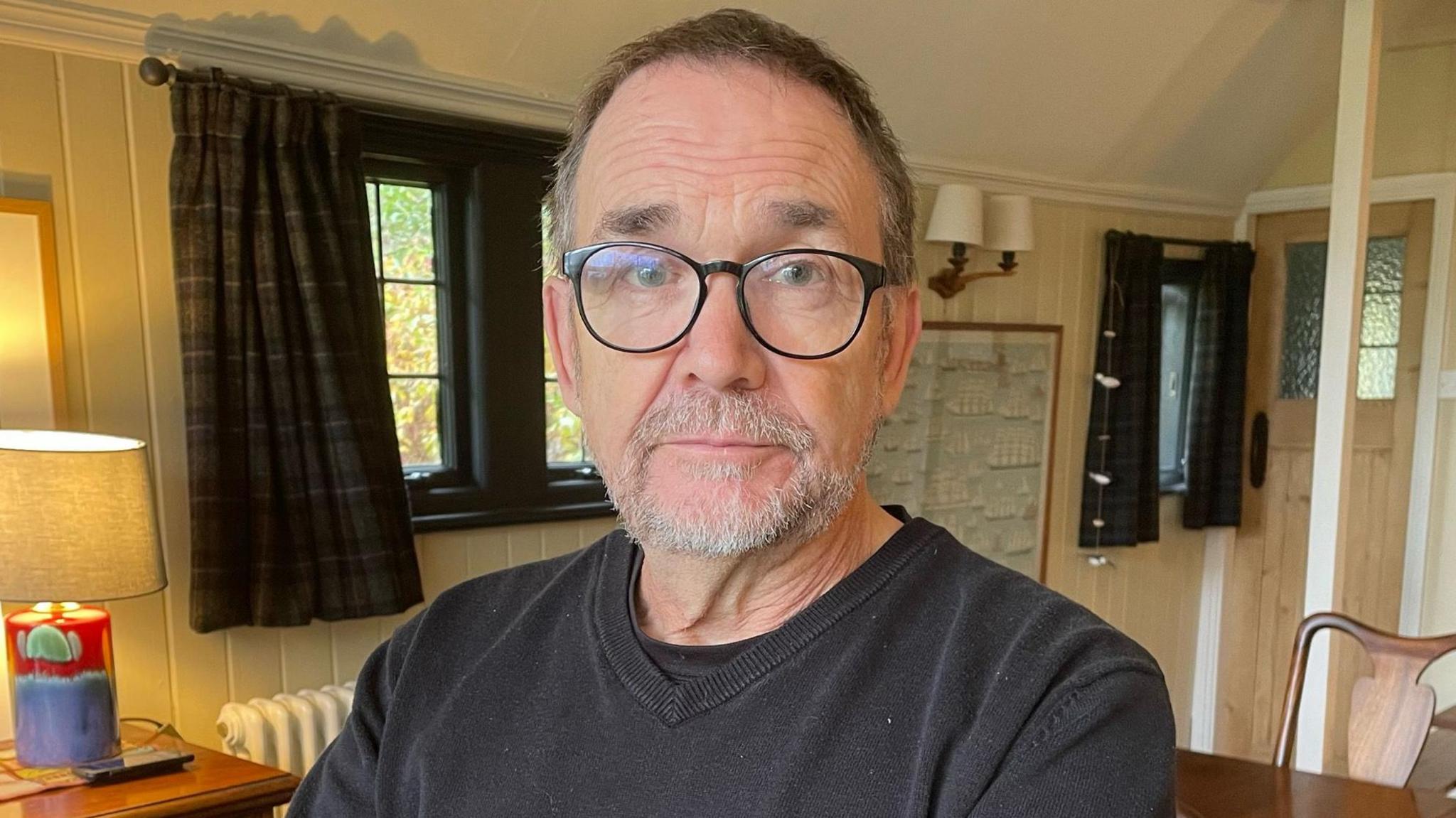 Jo Edye who has short grey cropped hair and a cropped white beard and is wearing round dark framed glasses and a grey sweatshirt. He is standing in his living room with a chest of drawers to his left and a table and chairs behind