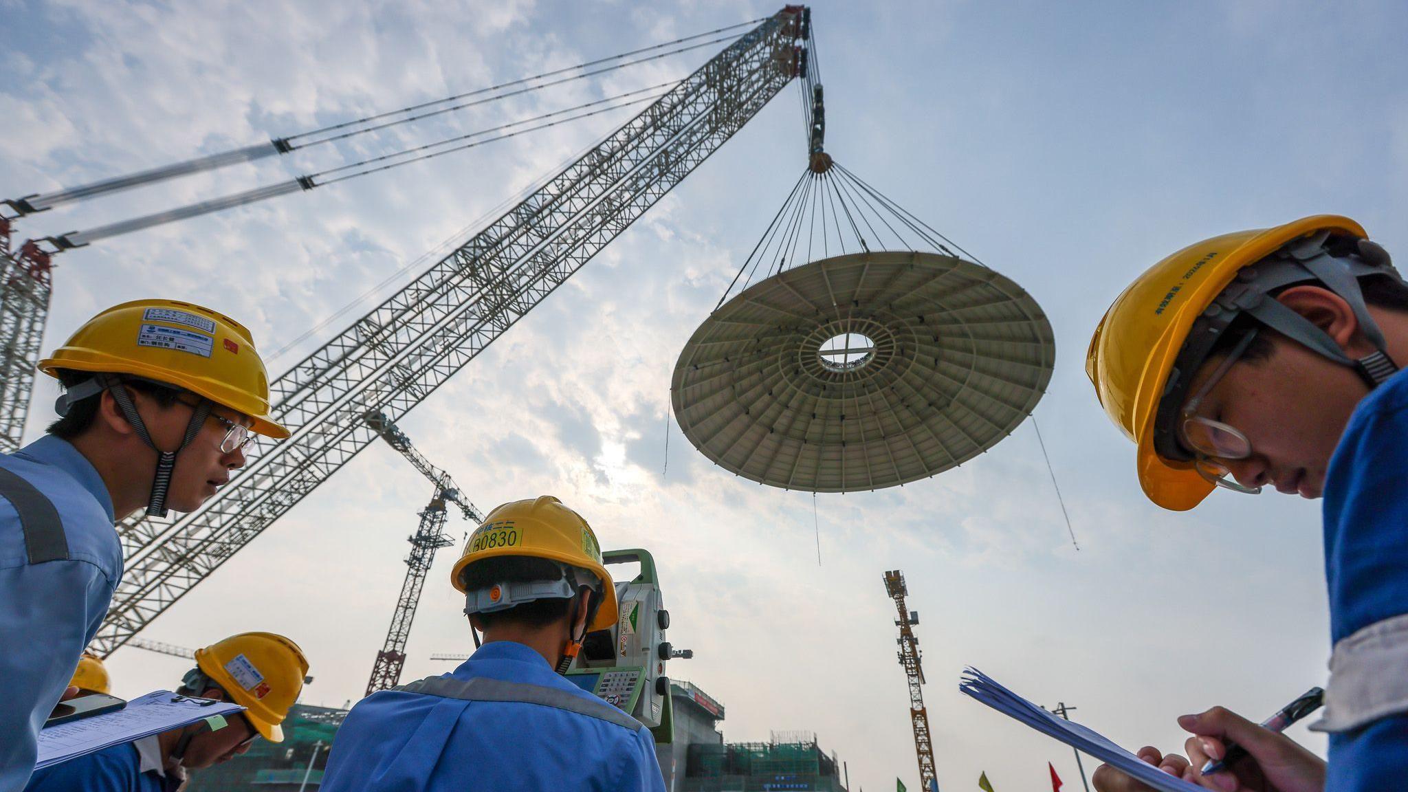 Men with hard hats watch as a small dome is lowered by giant cranes on the construction site of the world's first commercial SMR Linglong One in the Hainan Province of China in February 2024.