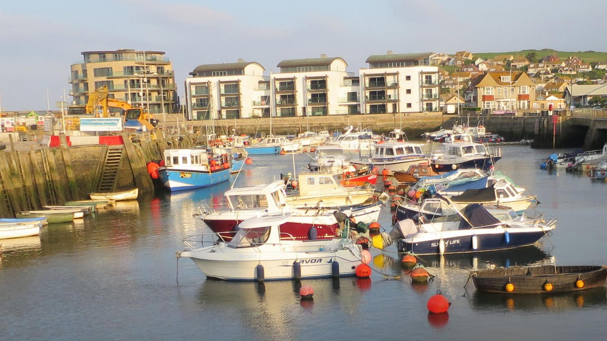West Bay harbour bathed in low sun. Dozens of boats are moored on the water which is surrounded by high harbour walls. Three modern white apartment blocks line the far end of the harbour wall. Another block can be seen off to the left and scores of houses can be seen on a hillside on the right of the picture.