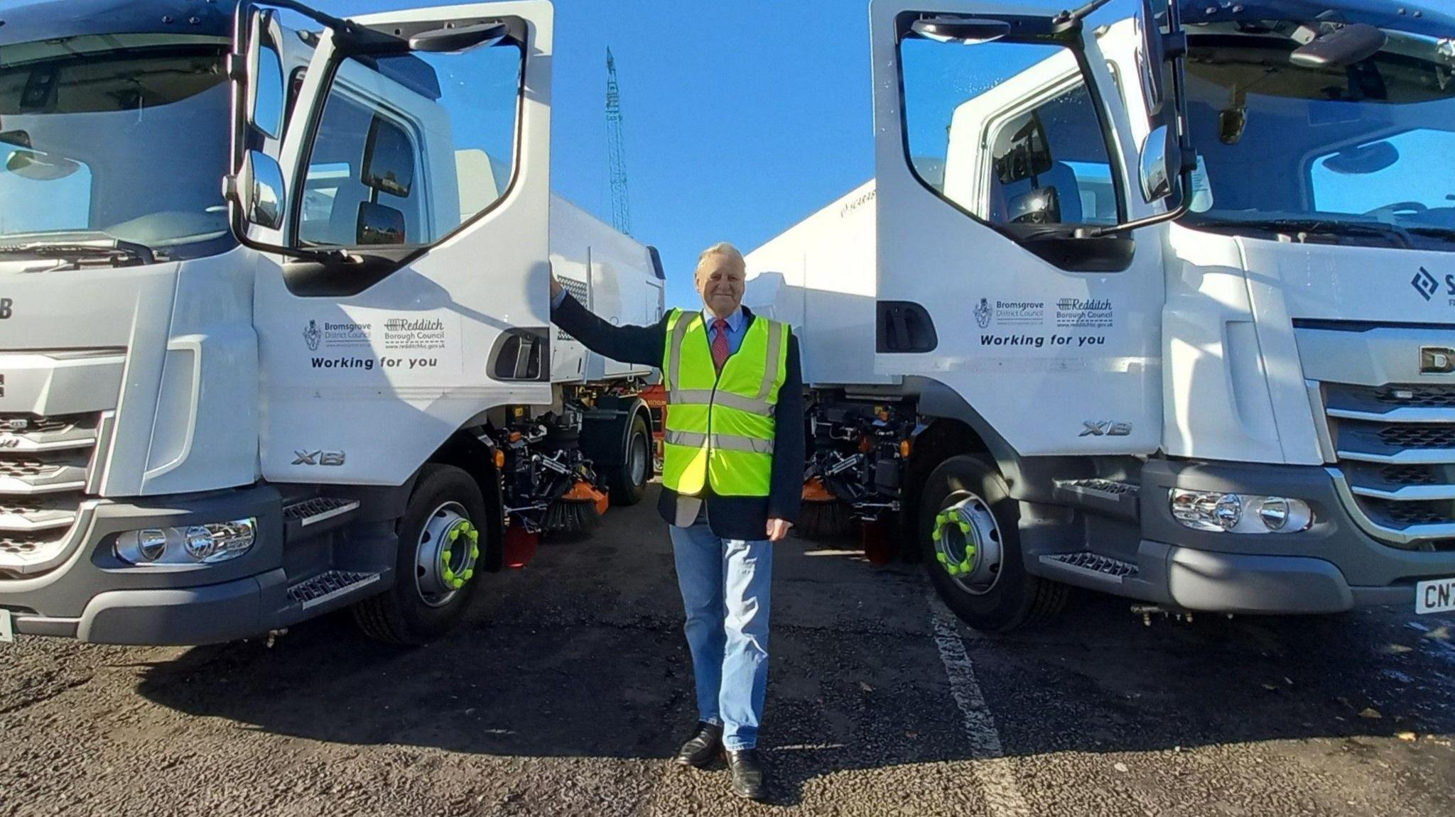 A man in a high viz jacket stands next to two large white trucks.