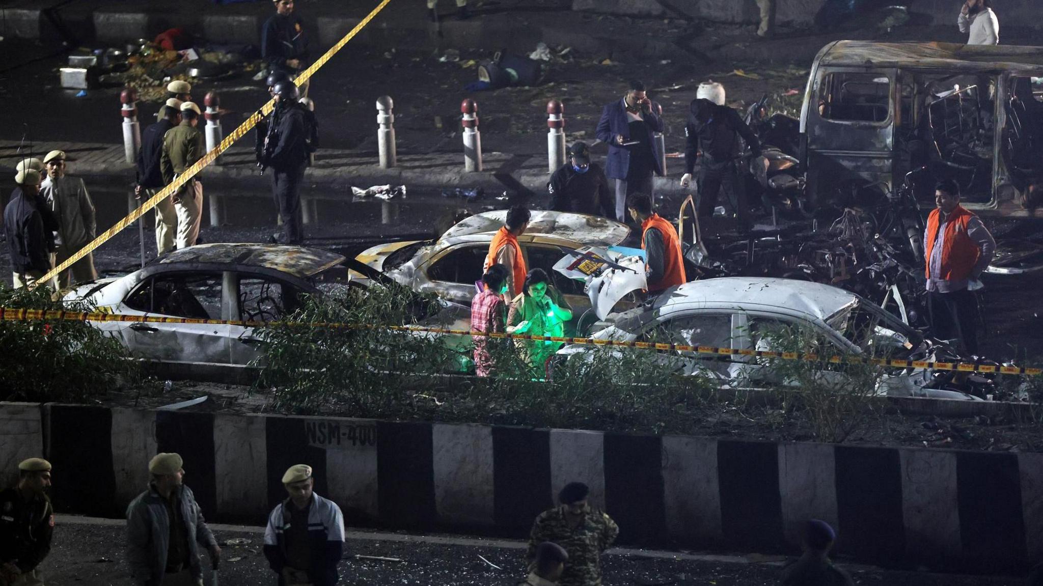 Indian police personnel inspect the scene of a blast at night. There's a few mangled vehicles, some with severe burn marks on them. Police tape can be seen marking off the scene in the top left corner of the image.