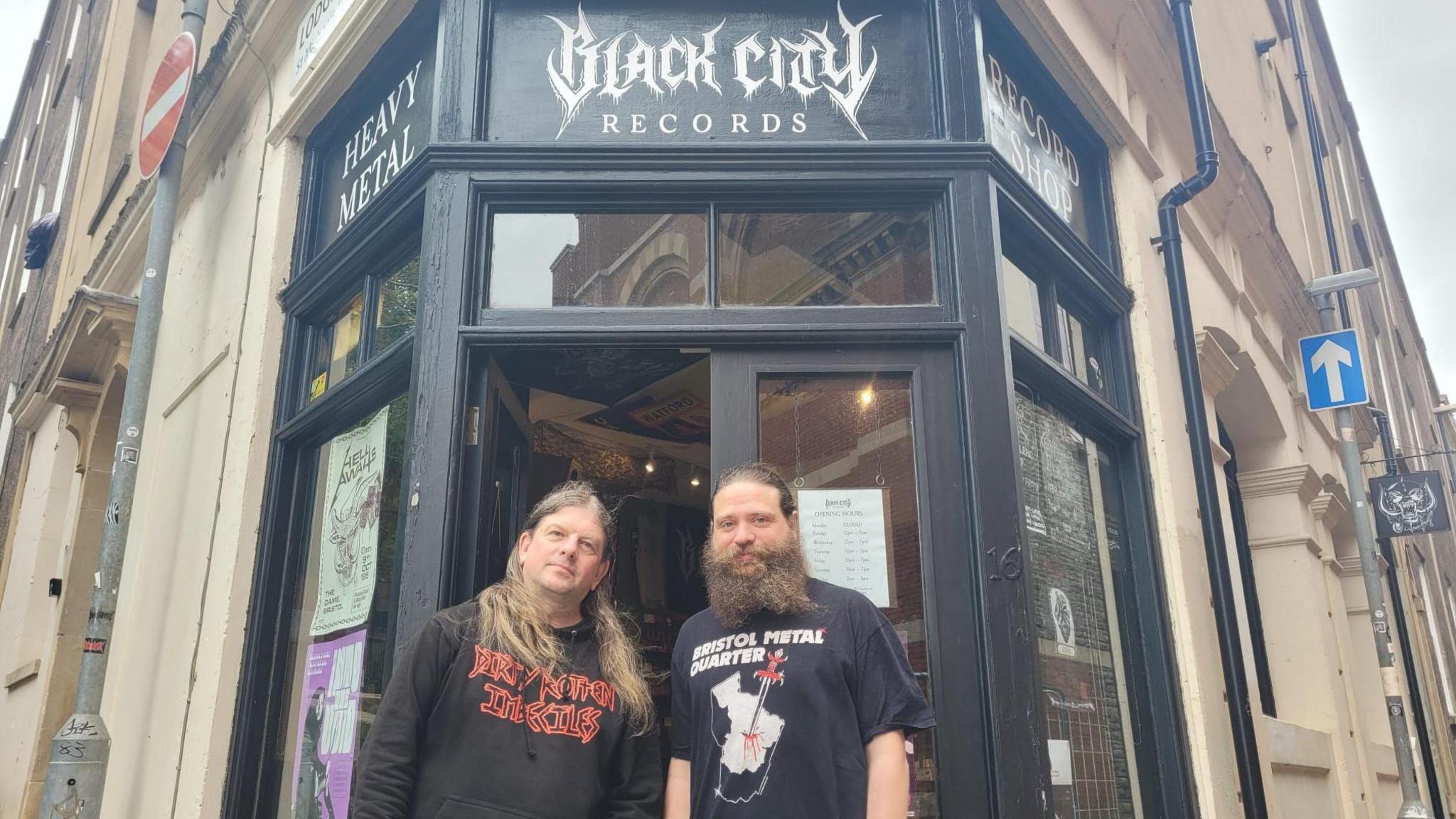 David Savage and John Ashby stand outside the entry to Black City Records. The shop is on the corner of a street and is painted black. 
