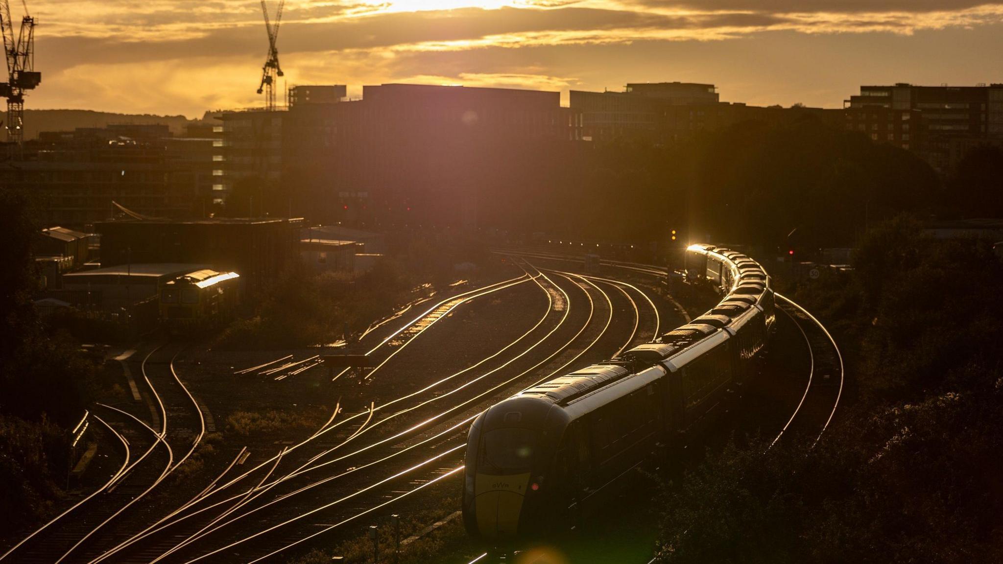 A Great Western Railway train approaches Bristol Temple Meads Station. The picture is taken at sunset with the tracks glowing bright in the setting sun and the sky tinged with orange. Buildings and cranes are silhouetted in the background