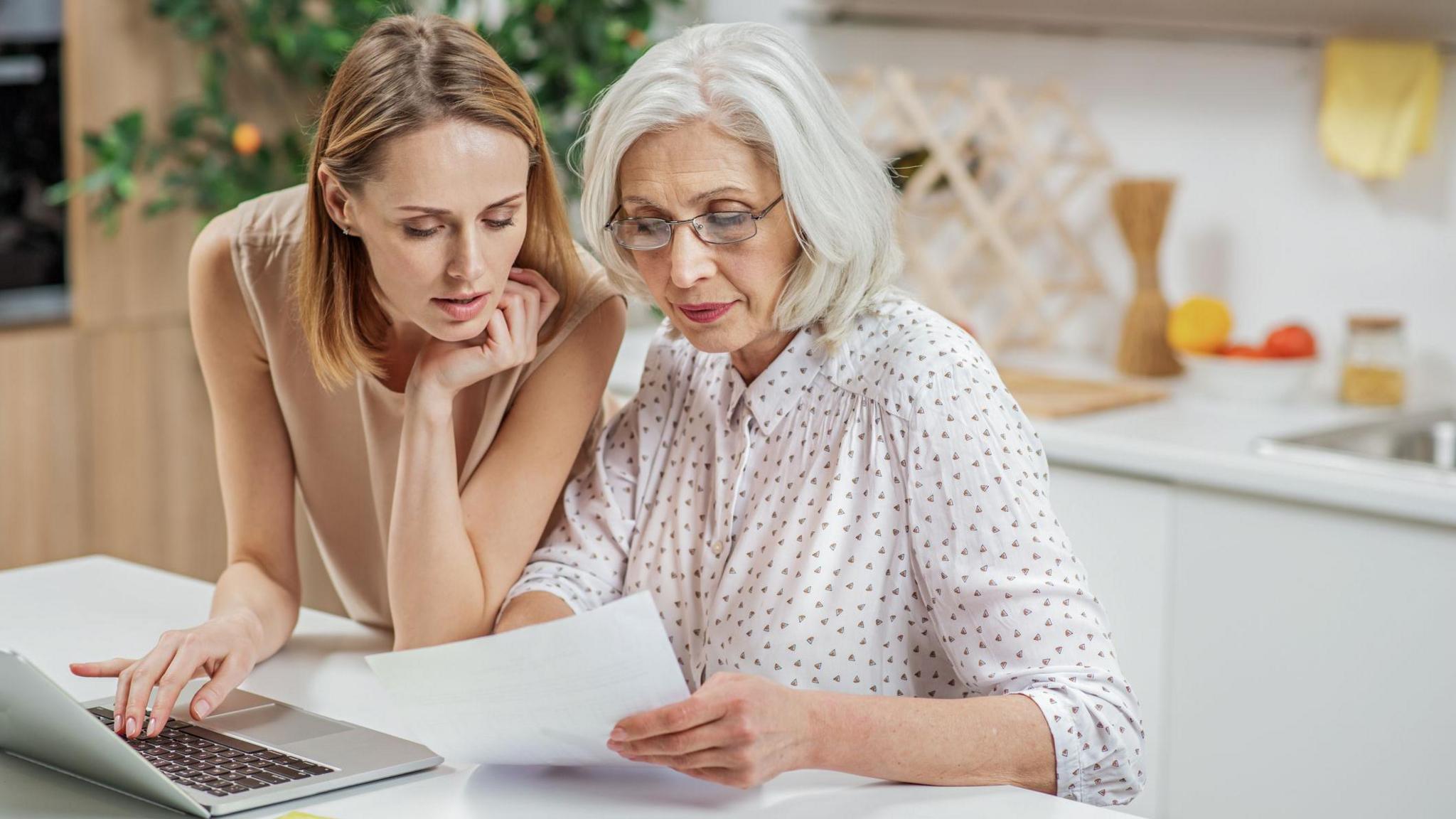 A young woman looks at paperwork with a woman who is a generation older. A laptop is open in front of them.