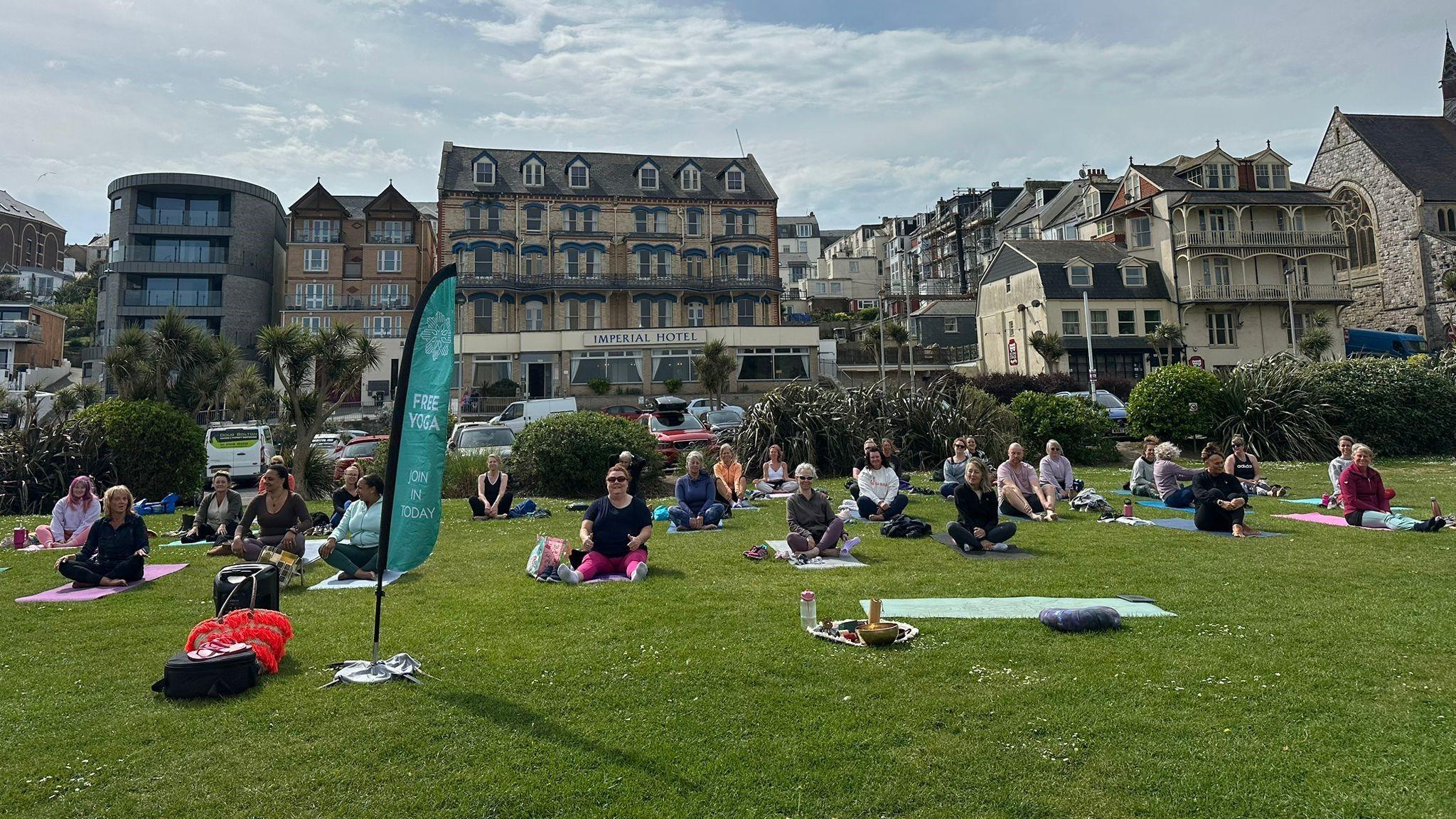 People sit on yoga mats in lotus position facing the camera. In the background, is the Imperial Hotel in Ilfracombe.