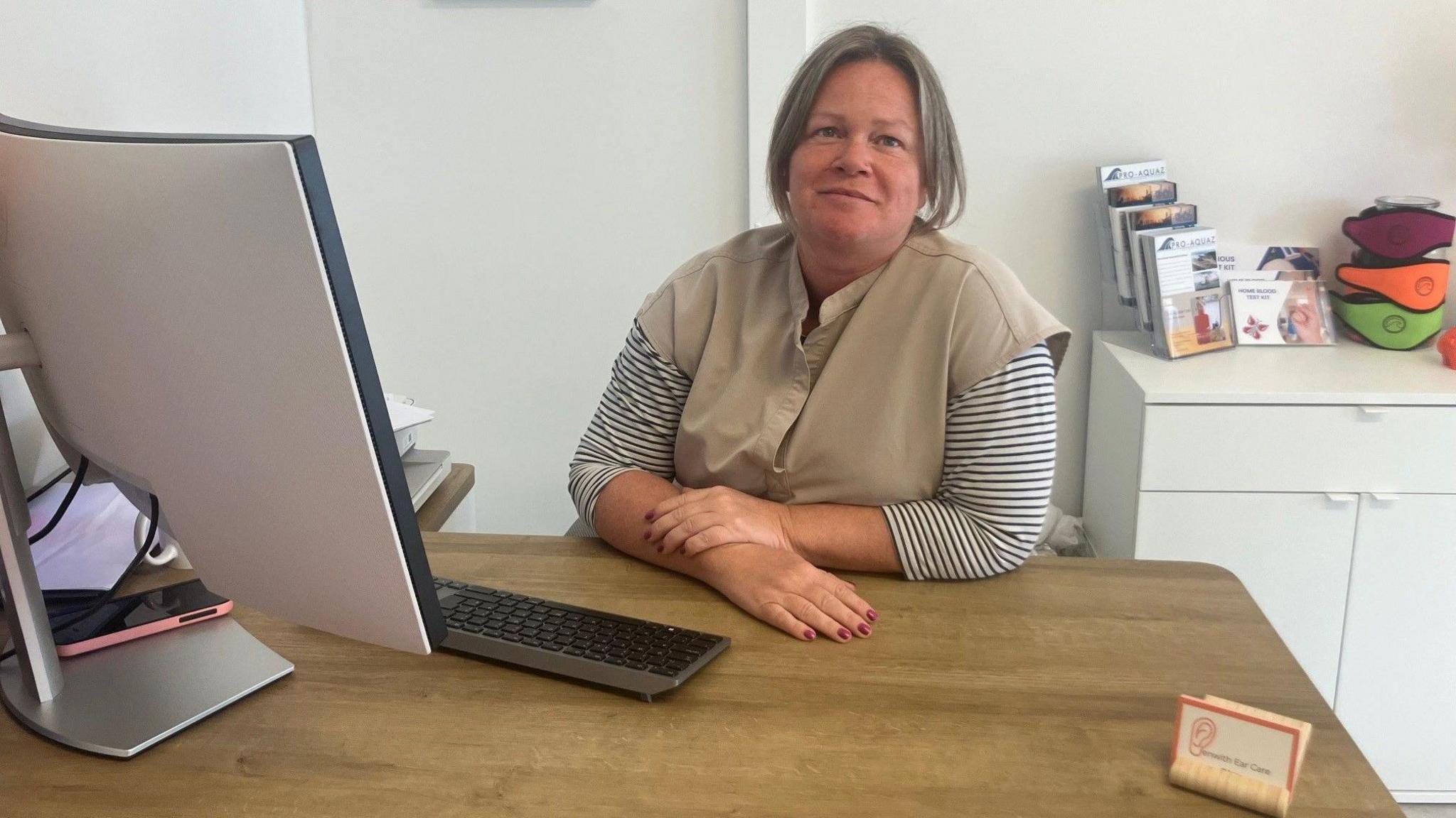 A woman sits at a desk with a computer. She runs an ear cleaning clinic , she wears a beige health workers tunic.