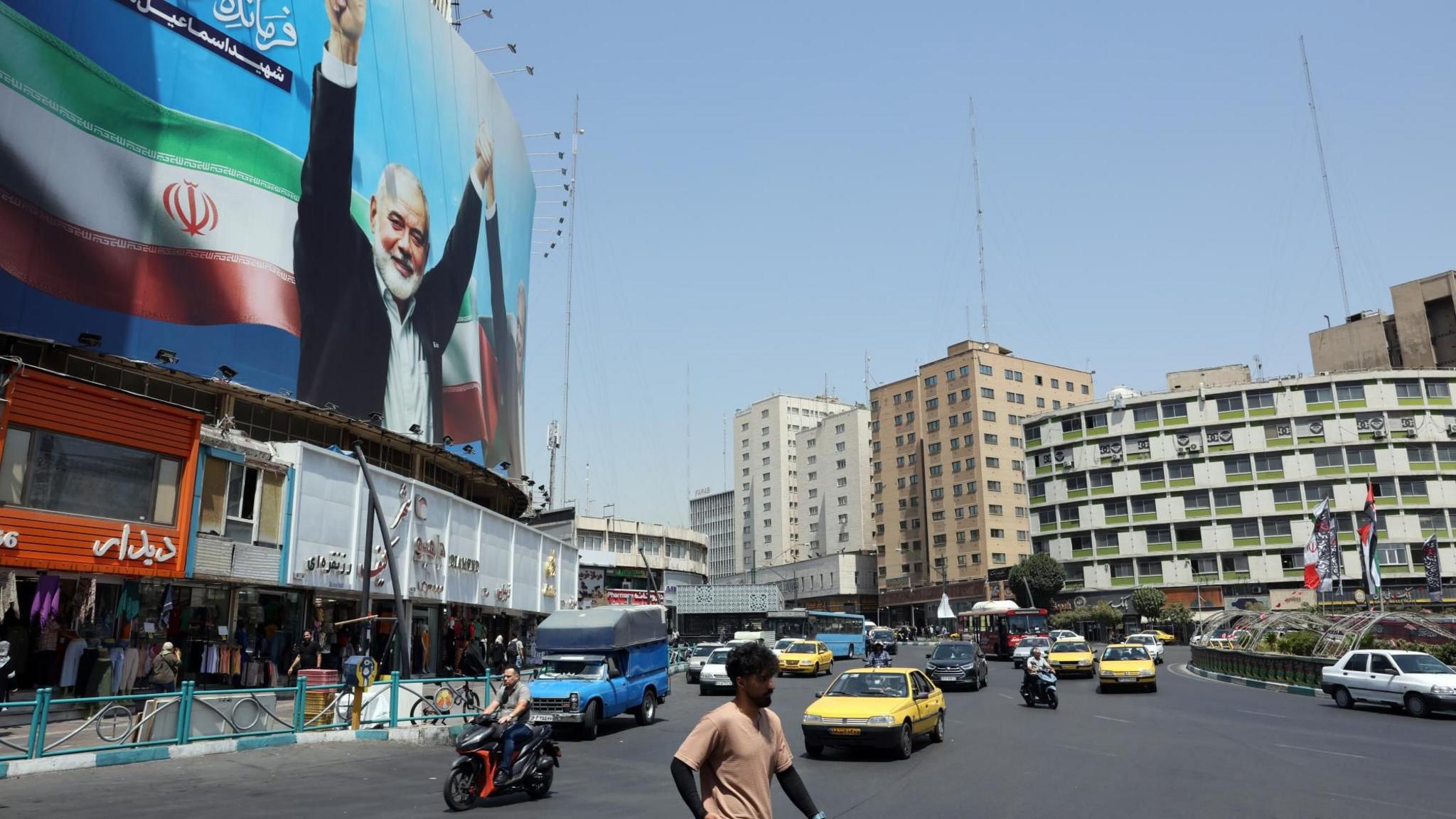 Iranians walk and drive past a billboard in Tehran, Iran, showing the late Hamas leader Ismail Haniyeh (13 August 2024)