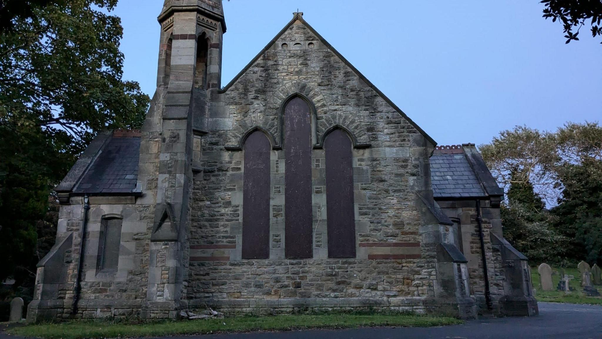 Ryton Chapel. The stone building's spire can be seen as well as three of its tall windows have been boarded up. Gravestones lie on either side of the building.