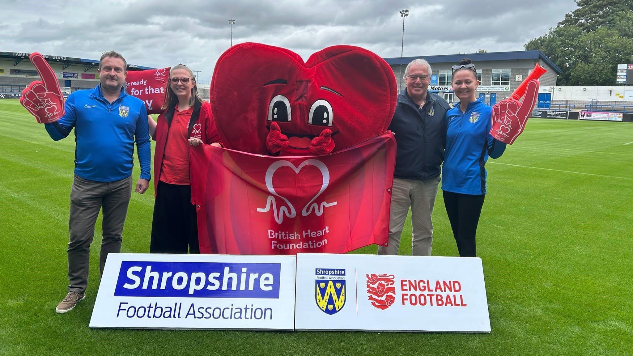 Four people are standing on a football pitch. In the middle is a person dressed as a giant red heart. There is a flag that reads "British Heart Foundation". Two of the people have red foam fingers on their hands.