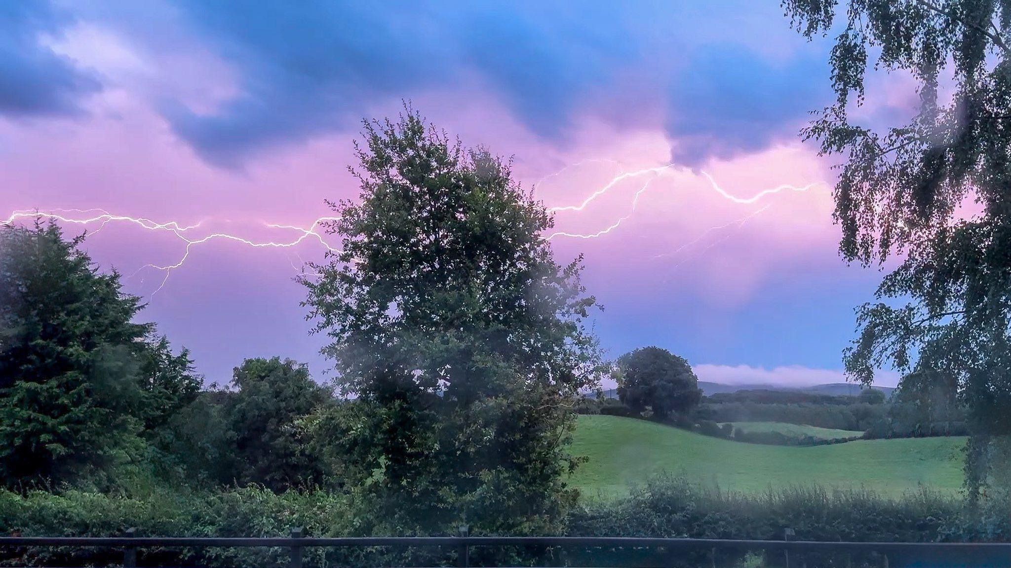 Lightning across the sky. The sky is a lilac and blue colour. There are trees and grass beneath it.