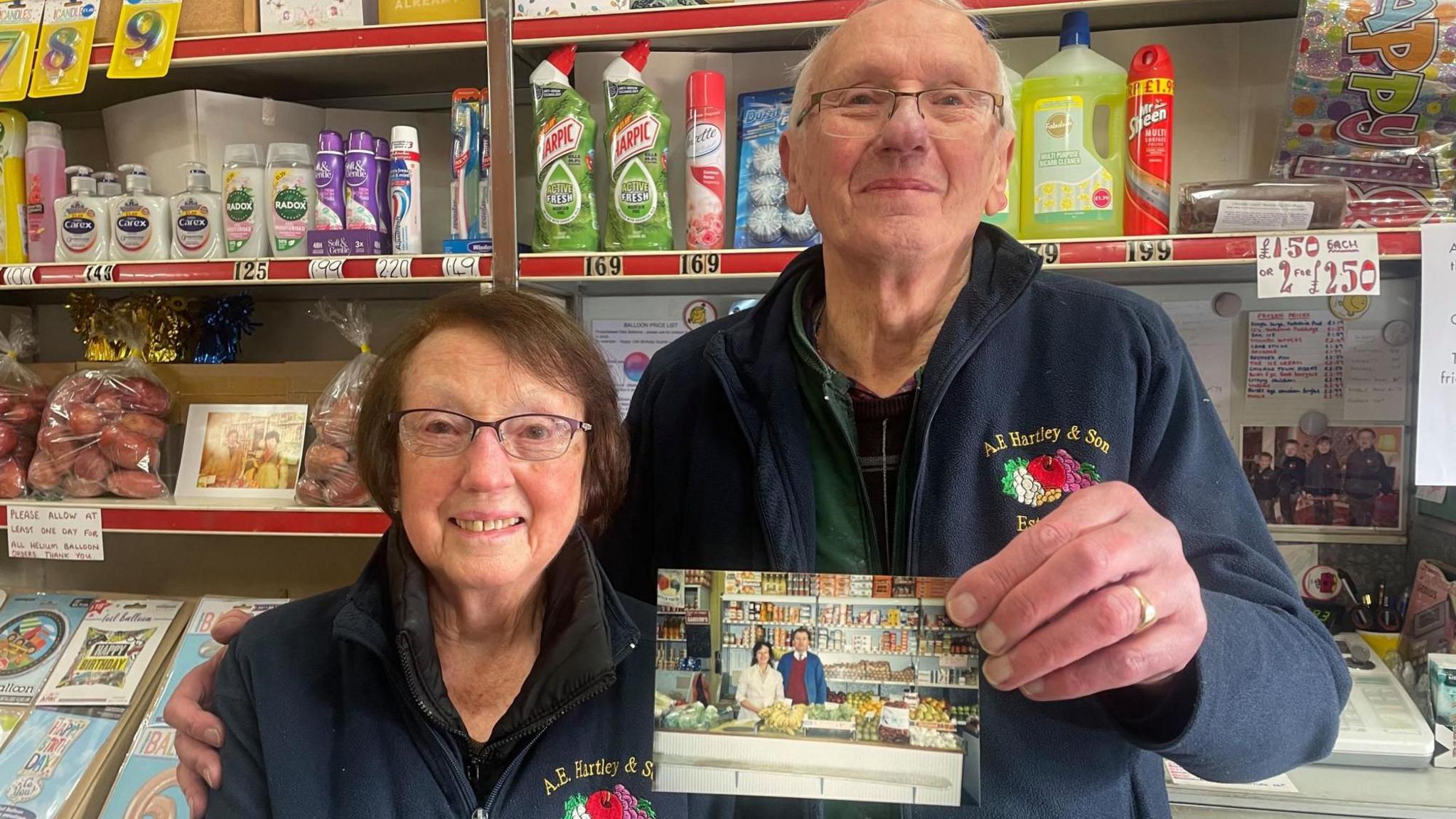 An elderly man and woman stand in a convenience store holding up an old photo of them when they were younger