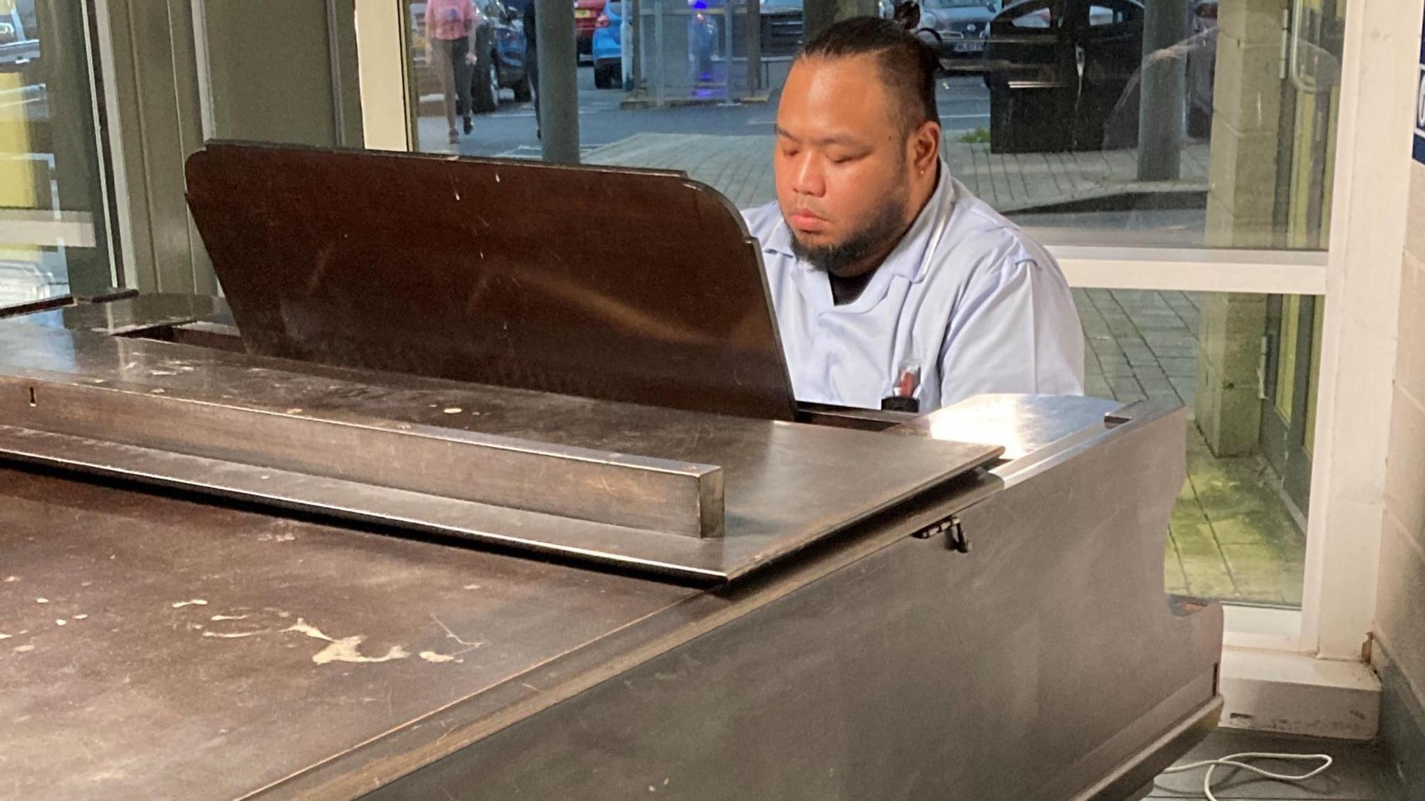 A man in a blue work uniform looking down as he plays at the piano. The piano is dark brown with some scratches on the top. 