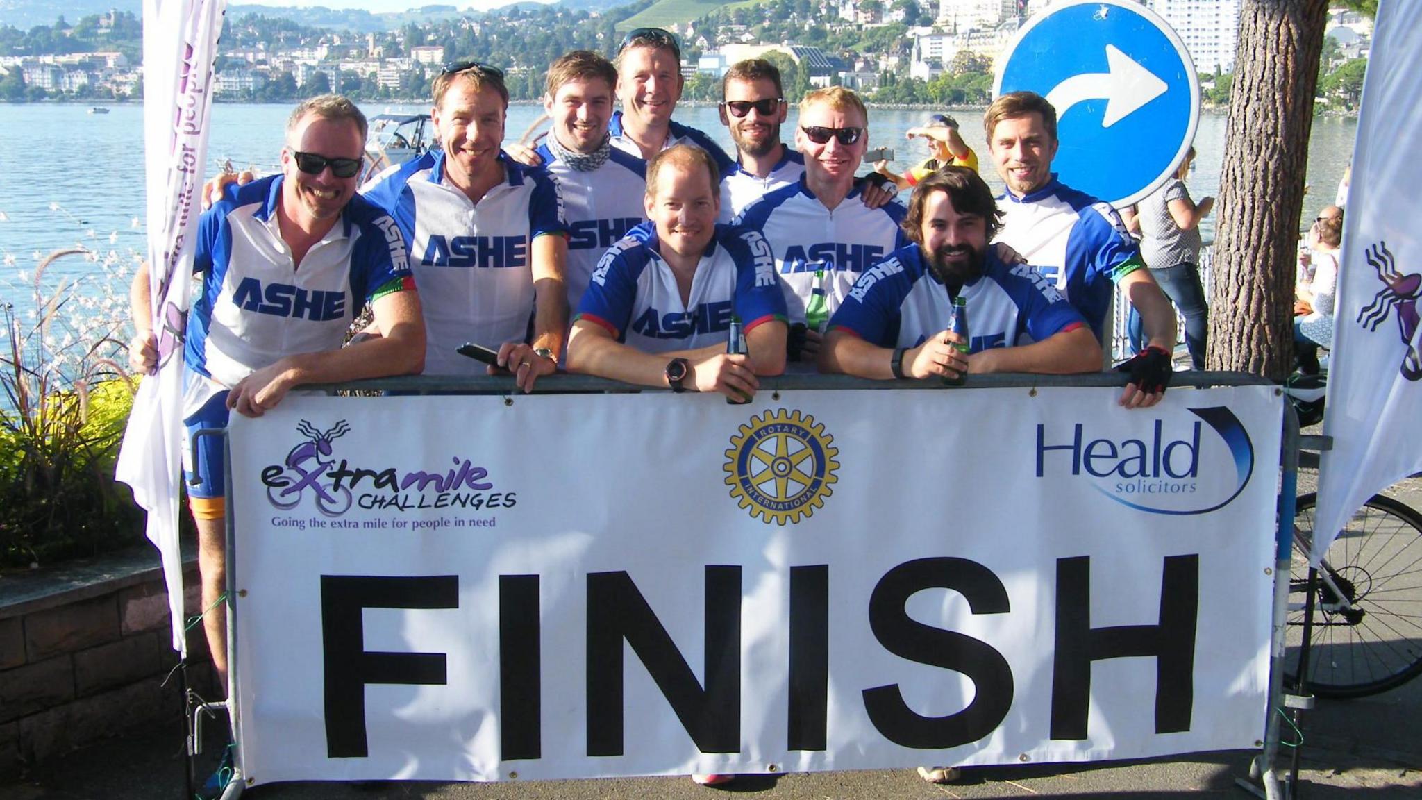 A large group of cyclists, standing by Lake Geneva, by a finish sign. There is a lake behind, with buildings and a sign. The group of men,  in white and blue tops are all looking at the camera and smiling. A few people are holding beers in the hands.