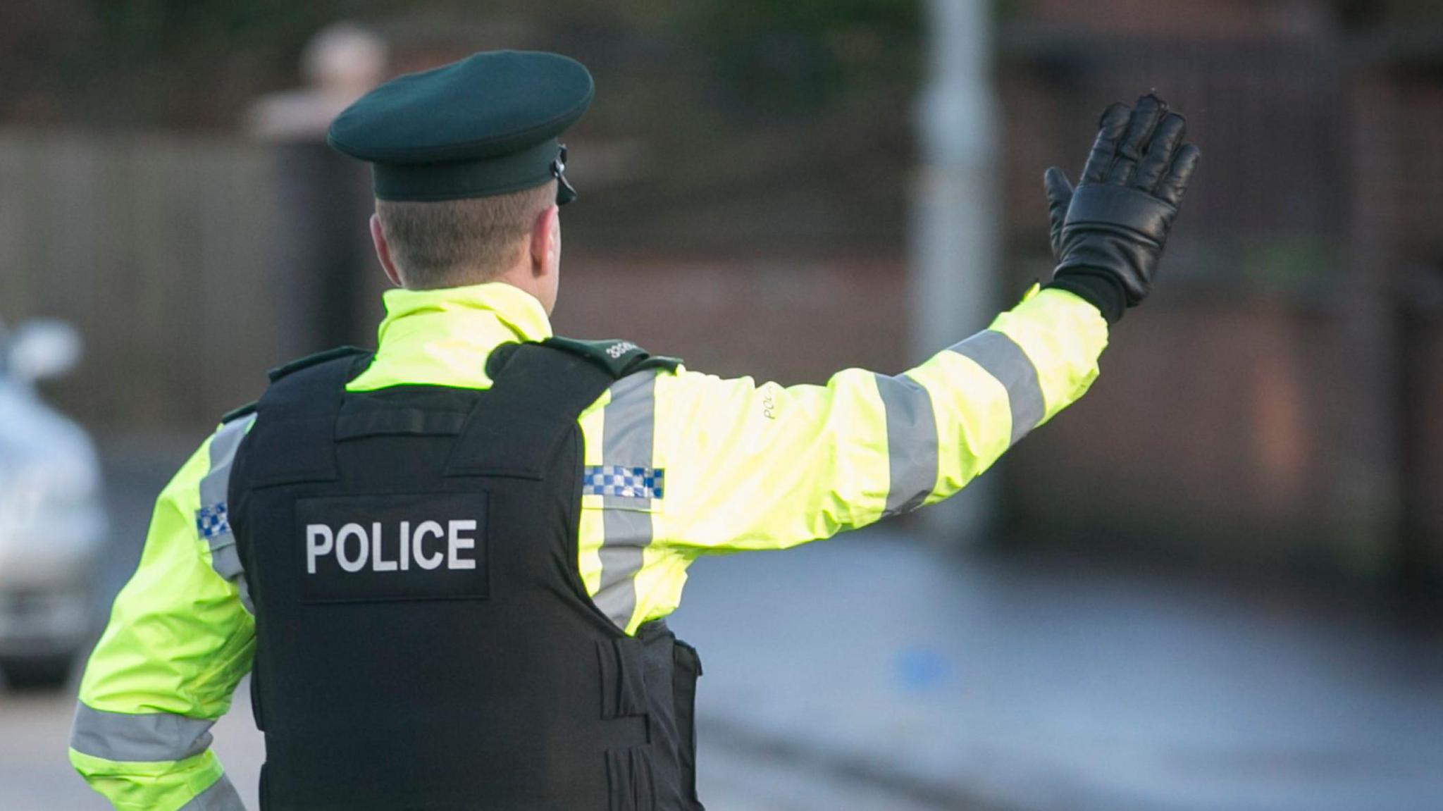 A PSNI officer is standing in the middle of a road wearing a protective vest, a green cap and a high-viz yellow jacket. His back is turned to the camera, has one hand on his hip and is holding the other out to flag down a car.