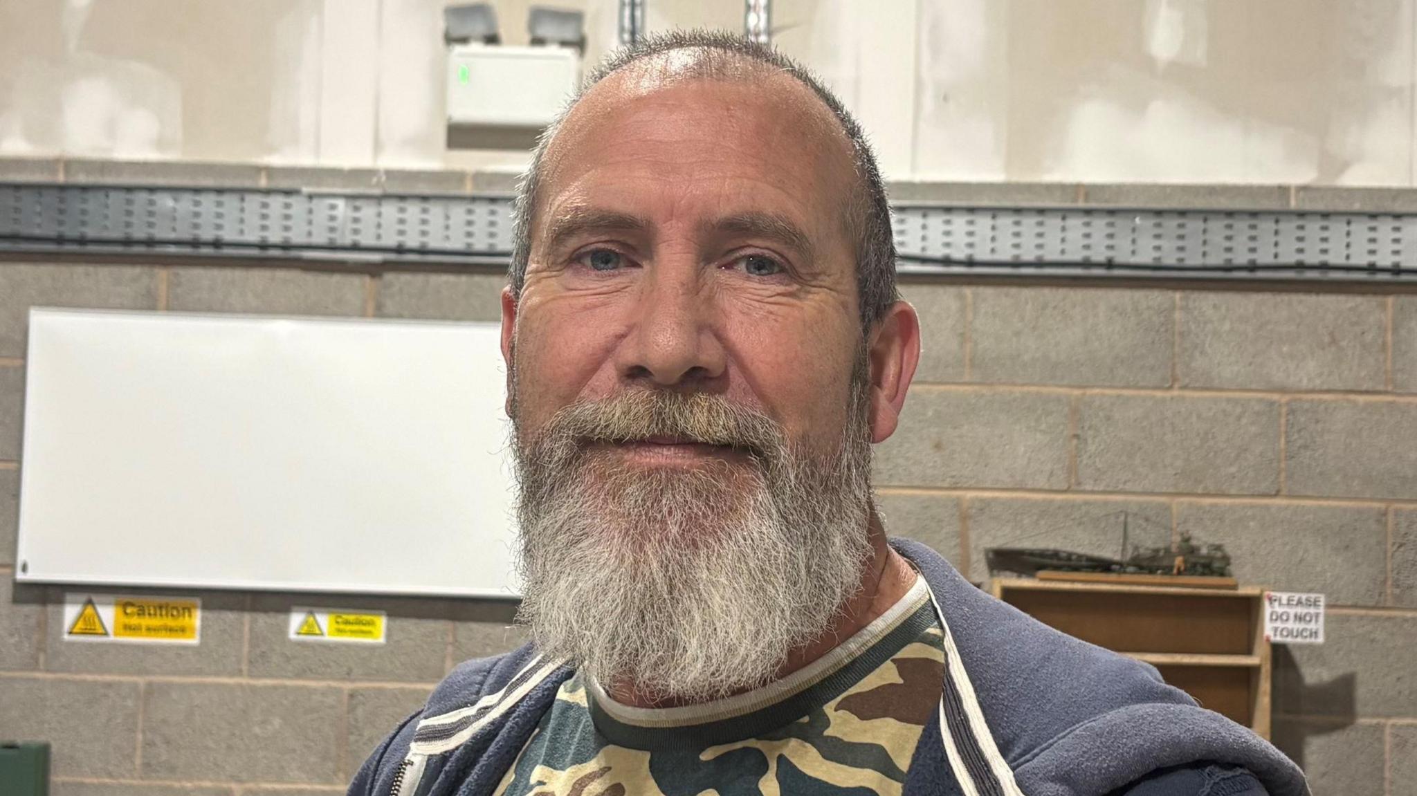A head and shoulders photo of Kurt Walton smiling at the camera. He has very short dark hair and a long grey beard. He is wearing a camoflauged t-shirt and a blue zip-up hoodie.