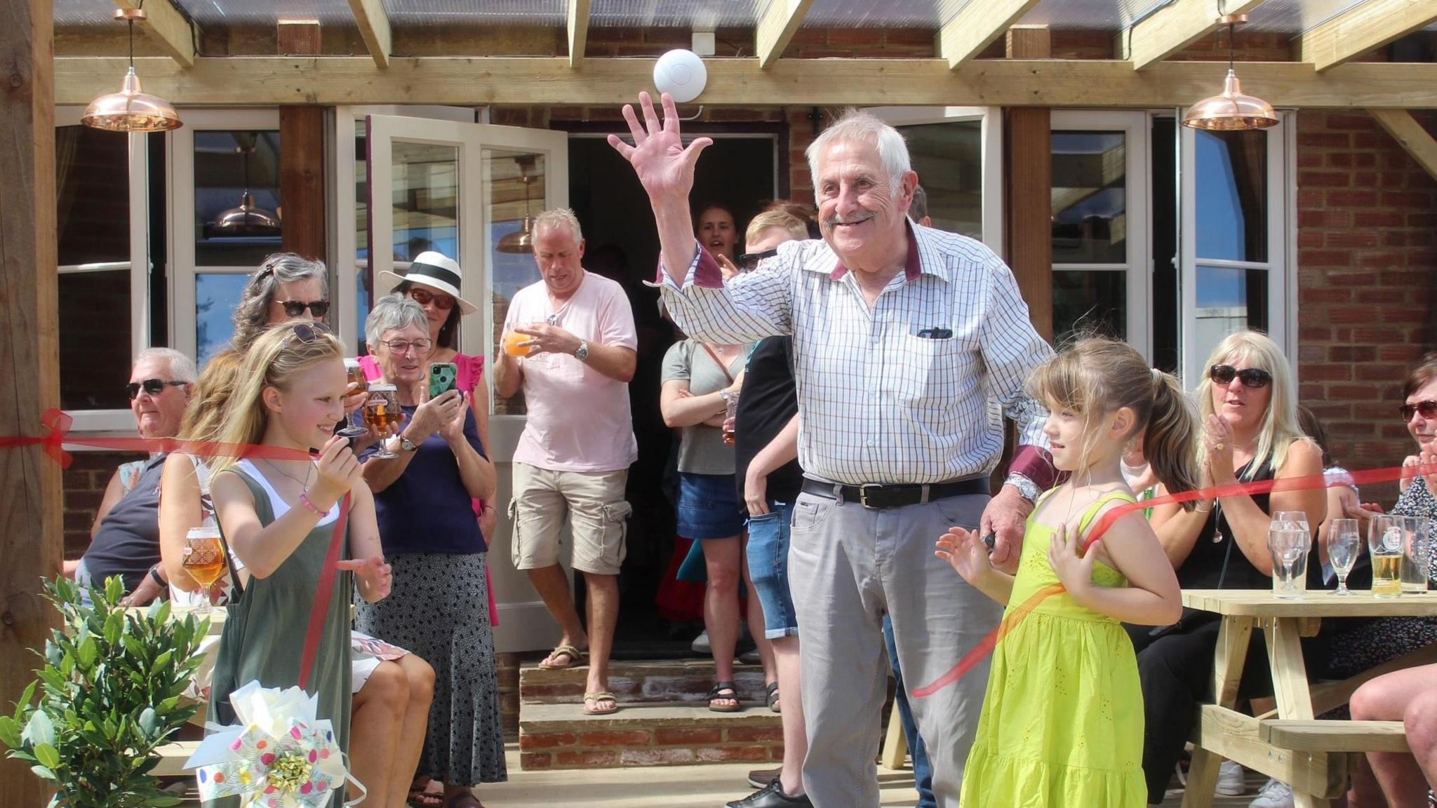 John Wicks stands smiling and waving as two young girls hold up the two sides of the recently cut ribbon. People sitting a standing in a crowd behind them smile and clap. 