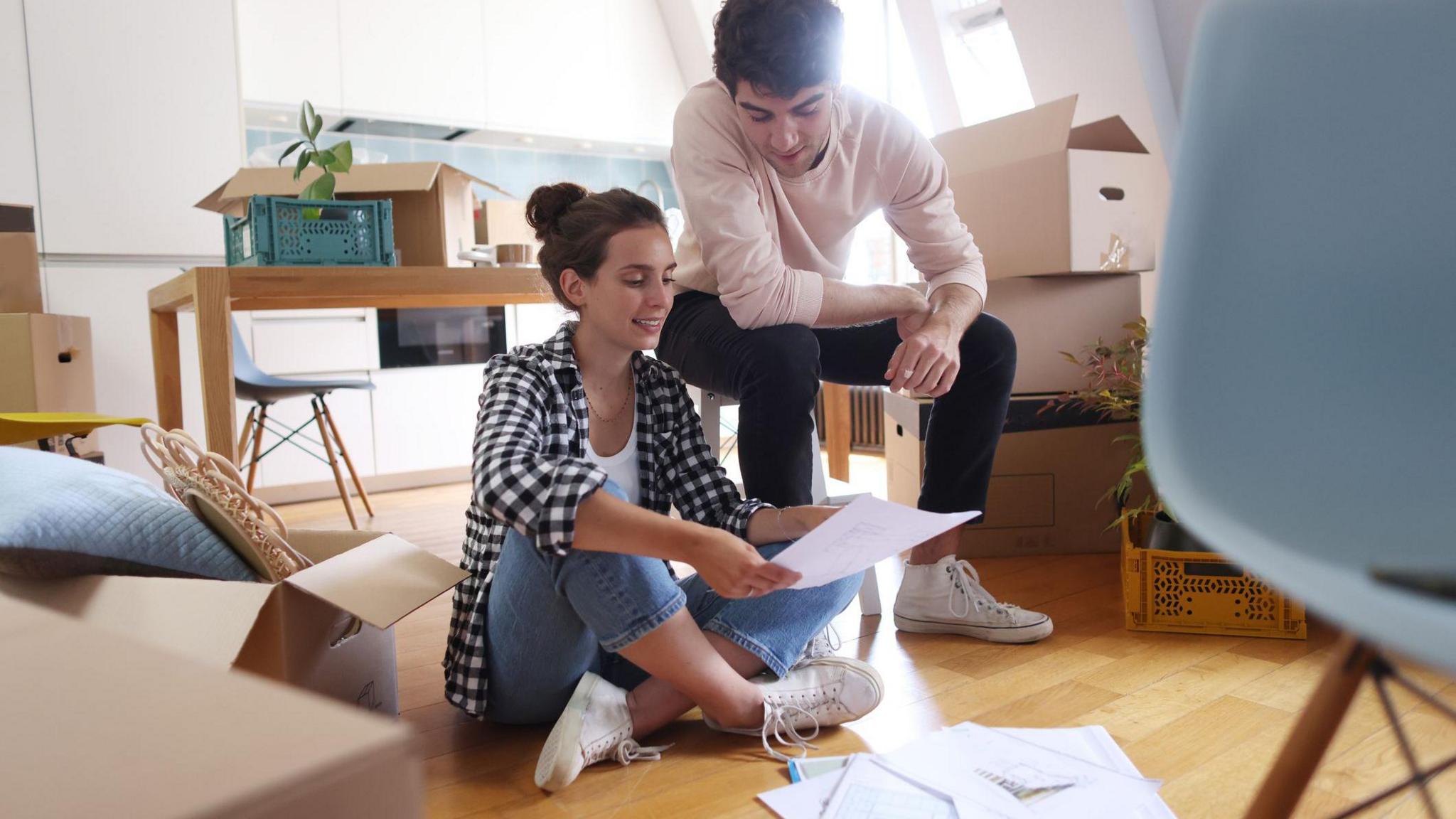 Casually-dressed man and a woman sit in a flat surrounded by cardboard boxes, as they look at paperwork