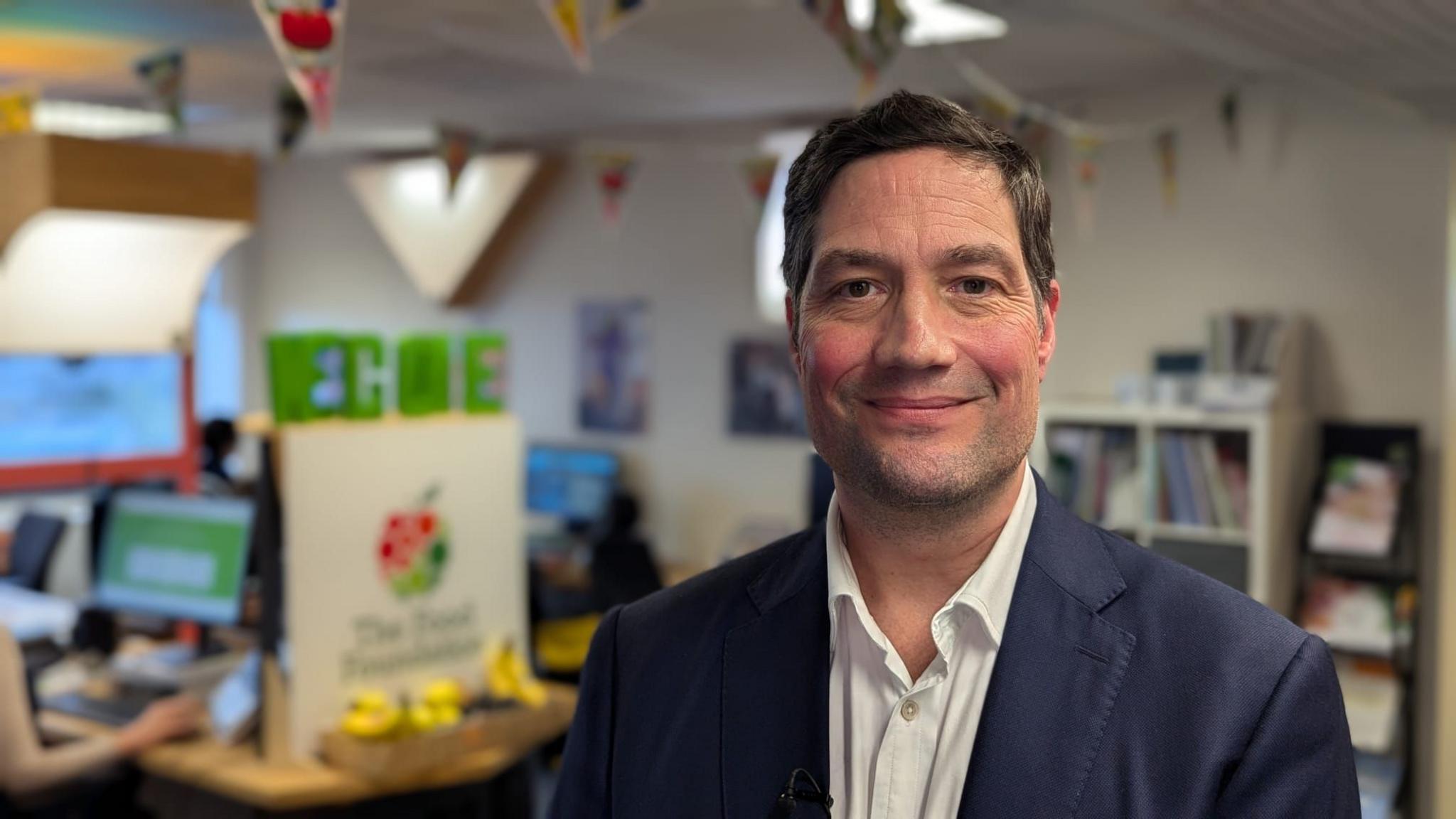 Myles Bremner smiles into the camera in a head and shoulders shot. He is wearing a navy suit jacket over an open-collared white shirt. He stands in front of a blurred office background with staff working on desktop computers.