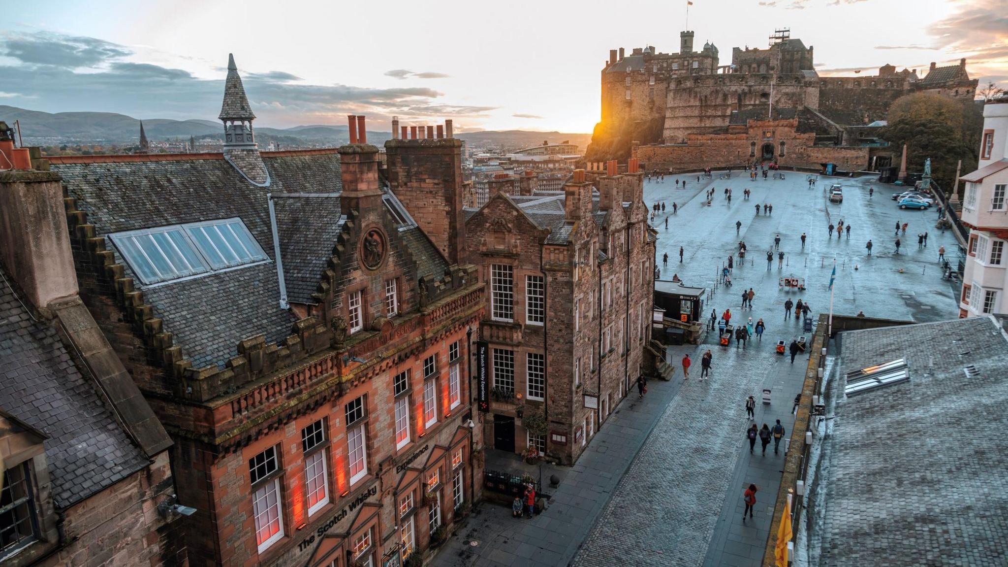 A rooftop view of Edinburgh Castle glowing in the warm colours of a sunset during the autumn season. Below the castle people walk along the esplanade 