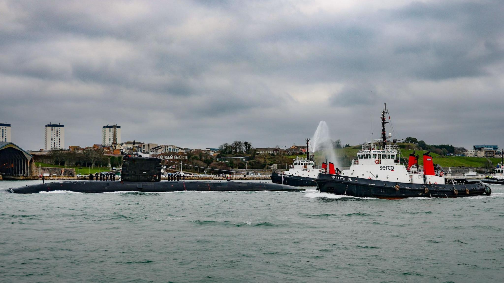 The submarine arriving at port, accompanied by two tugboats firing water cannons. The skies above are grey and overcast