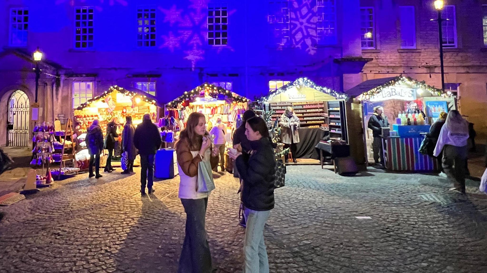 People gathered outside chalets on an evening at Bath Christmas Market, with Christmas lights on the chalets and the buildings behind illuminated in blue