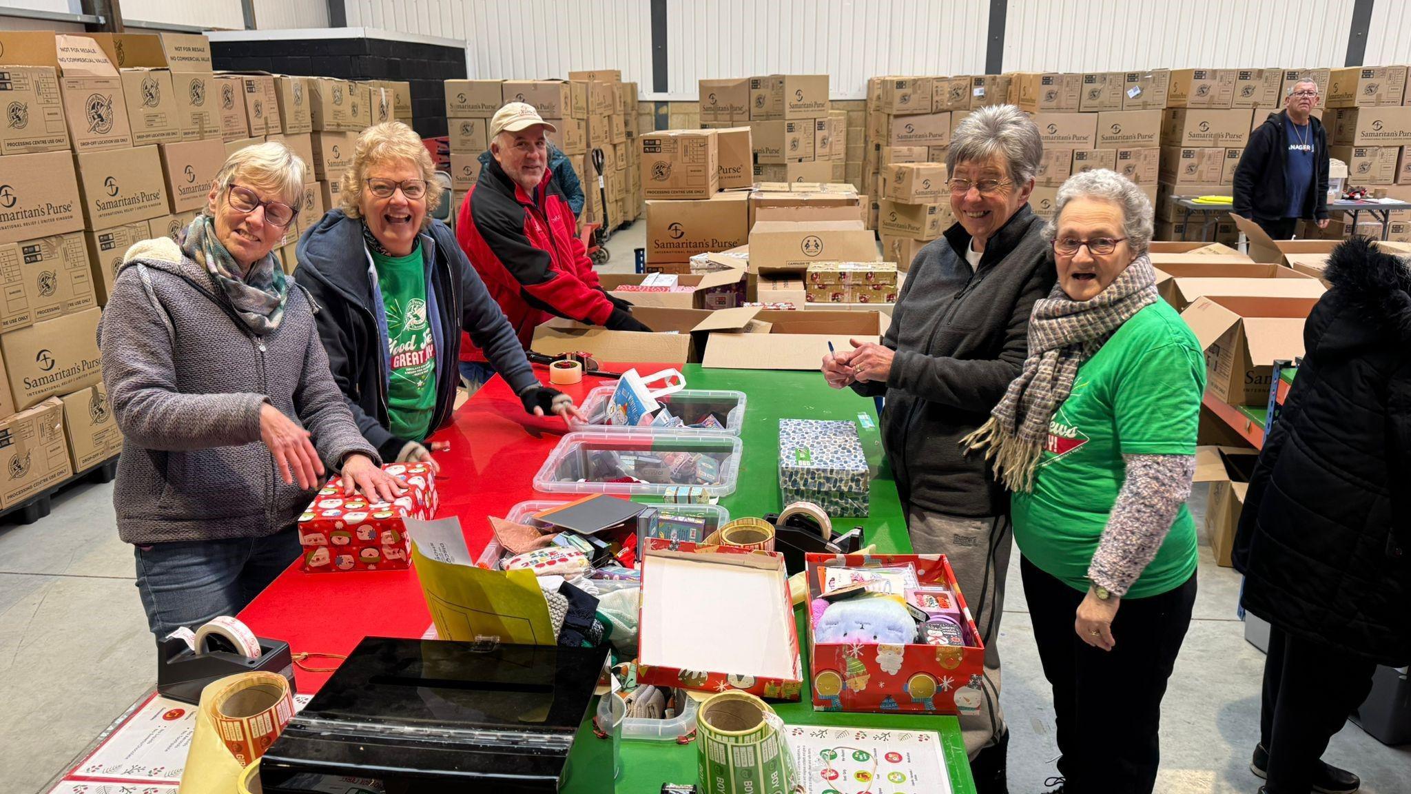 Five volunteers smiling at the camera. They are standing around a table which has wrapped shoeboxes on. 