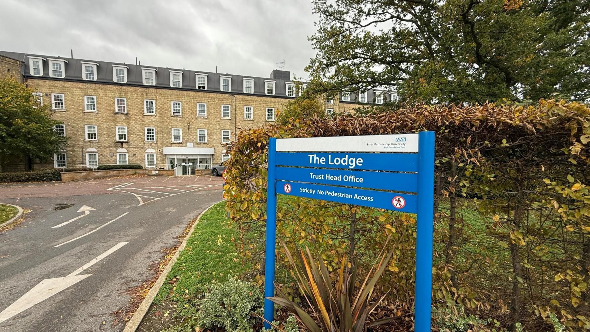 The Lodge in Essex is the headquarters of the Essex Partnership University NHS Foundation Trust and is shown in this picture. It is made up of light yellow bricks and is four storeys tall. It is a long building with trees to the front of it. There is a car in the immediate foreground in the car park. The window frames are white. There is an access road to the front of the picture.