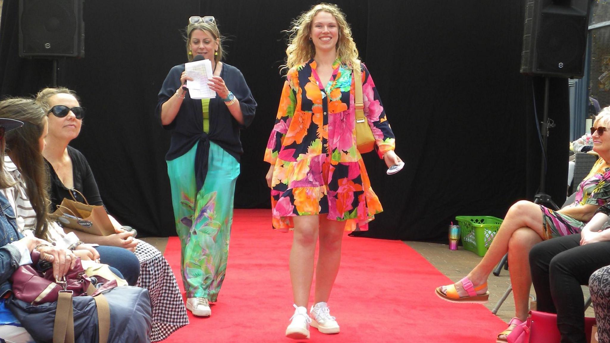 Woman with long blond hair wearing a multi-coloured dress in light fabric and an orange handbag. She also has white plimsolls. She is walking down a red carpet. There is a woman behind her wearing a black T-shirt and blue trousers with a floral design above white plimsolls, who is reading from a piece of white paper into a microphone. There are people sitting on either side of the carpet.