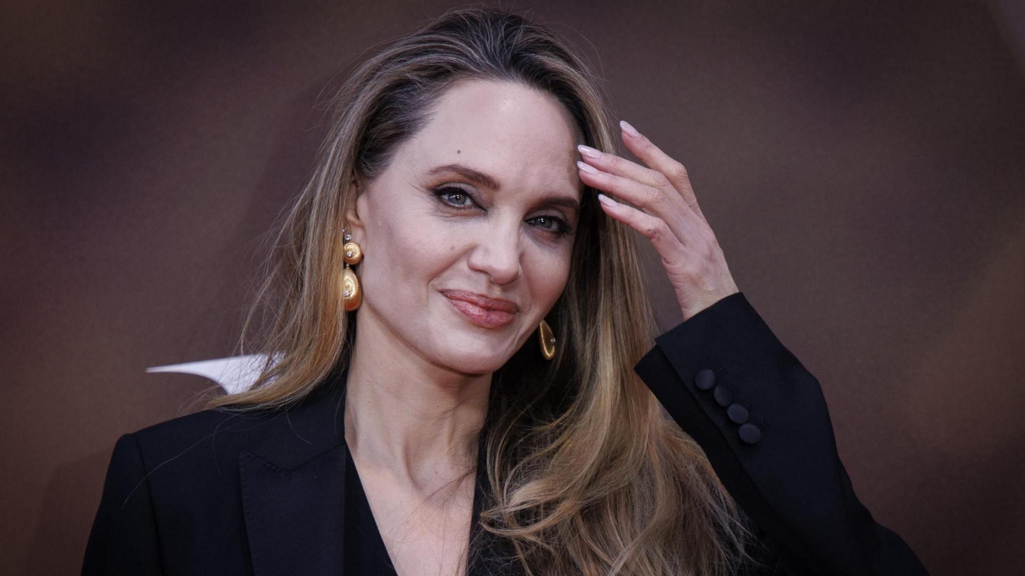 Angelina Jolie smiles at the camera. She has long, fair hair and is wearing gold dangly earrings. She is also wearing a black jacket and is touching her face. 