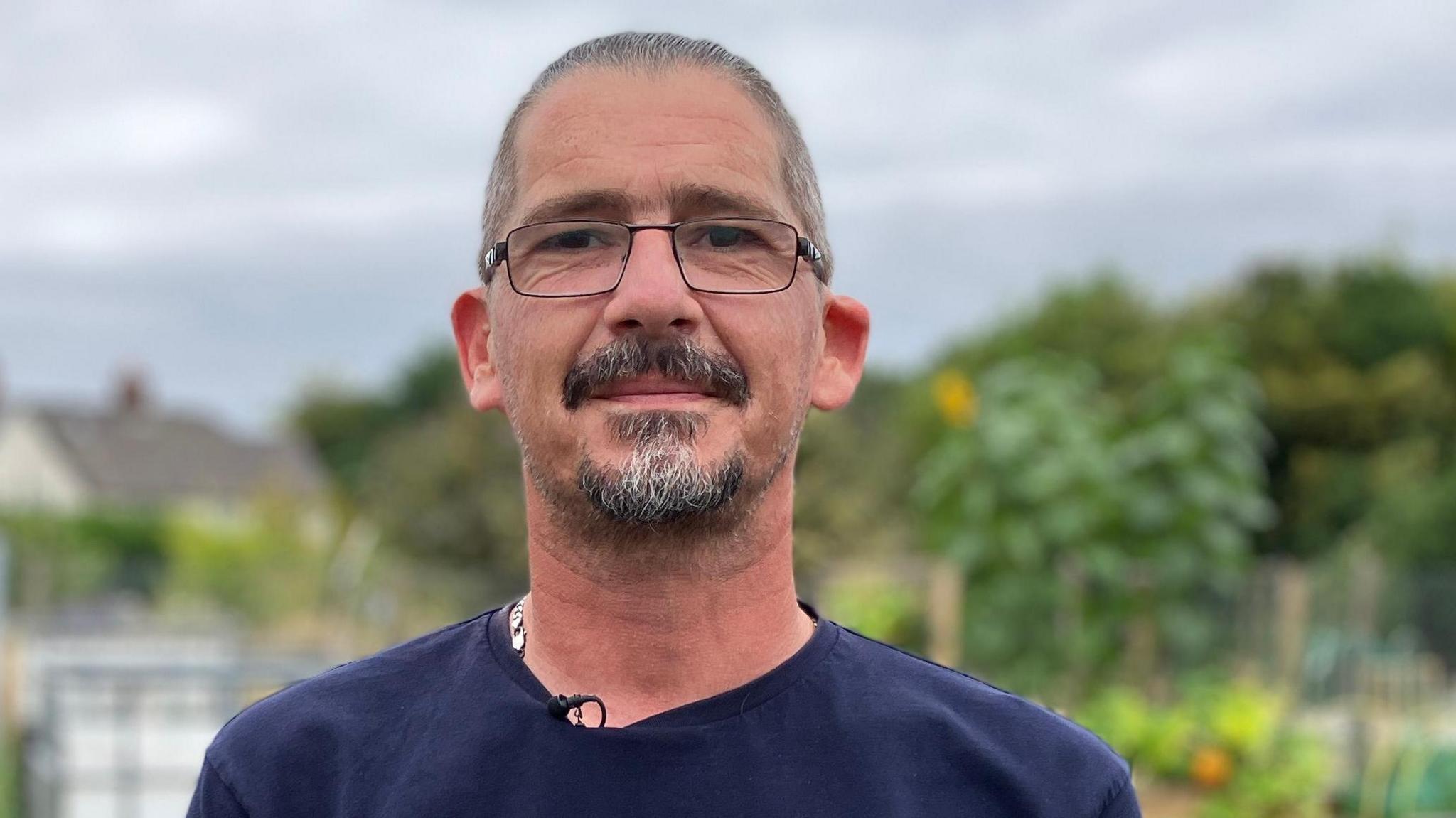 Ricky smiling at the camera. He has shaved grey hair, a moustache and goatee beard. He wears glasses and a navy blue t shirt. He is standing in an allotment with trees in the distance. He is in focus but the image behind him of the allotment is blurred.