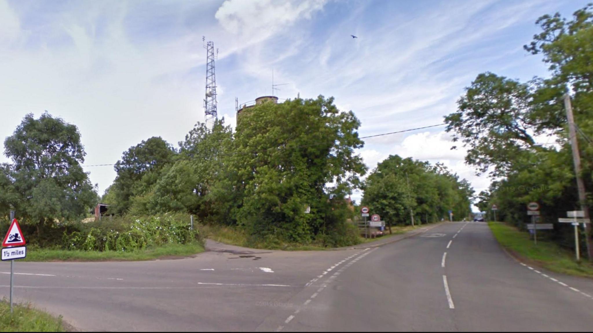 Rural road junction showing entrance to the village of Barby. Village signs are visible in the distance, and there is an old-fashioned white signpost to the right. The road is bordered by trees on both sides, and there is a left turn onto Longdown Lane which includes a humped-back bridge sign.