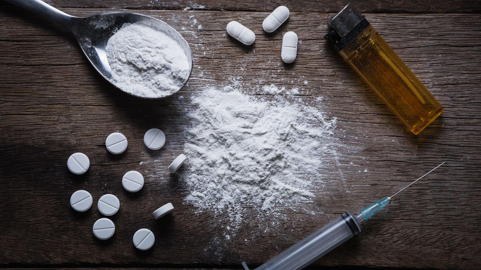 A brown table, on which several white pills are scattered alongside a lighter, a syringe and a spoon full of white powder. There is more white powder on the table.