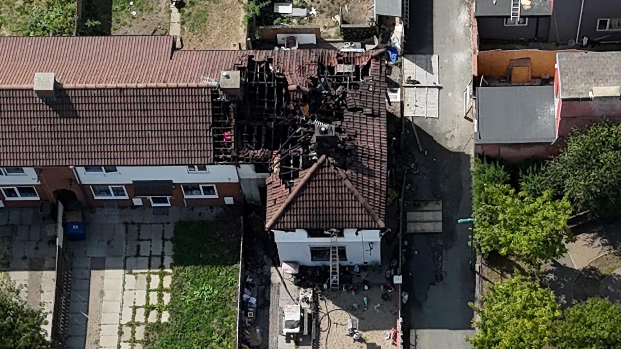 An aerial view of the damaged house. There is a large hole in the brown roof with tiles in disarray. A ladder is propped up against the white house. There are three other properties attached to it.
