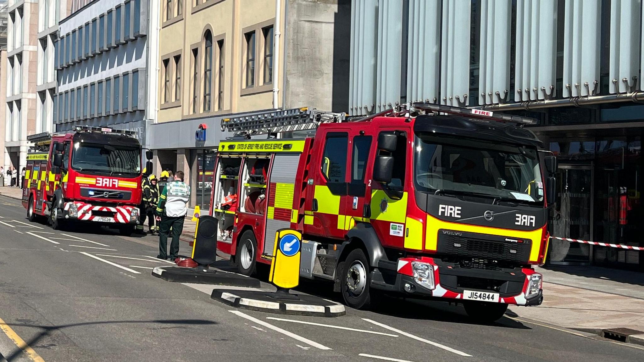 Two fire engines are parked next to buildings lining a road. There are a couple of firemen in uniform standing next to one of the engines. The engines are branded with red and fluorescent yellow checks.