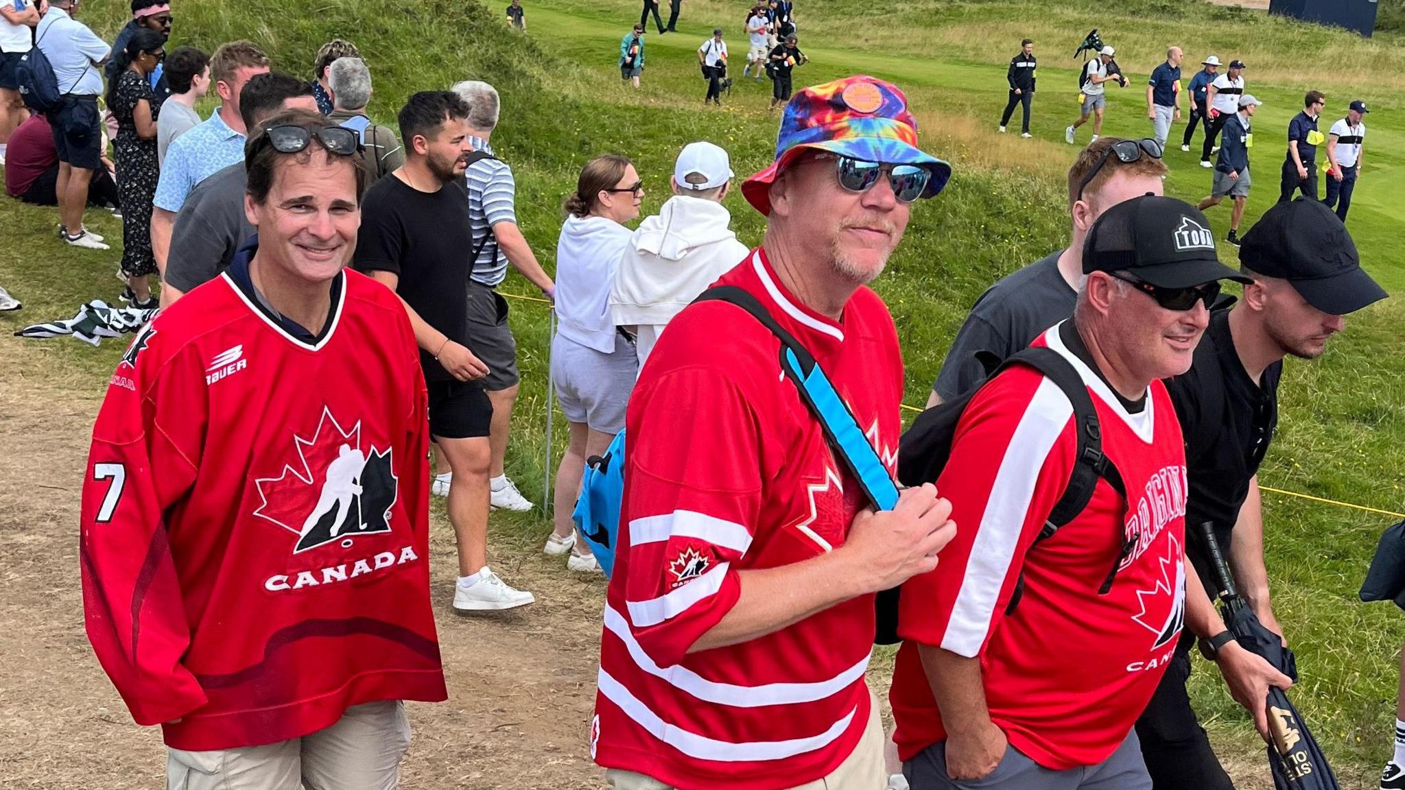 Three men wearing red Canadian jerseys. They are standing on the grounds of The Open at Royal Portrush, with a crowd of people in the background.