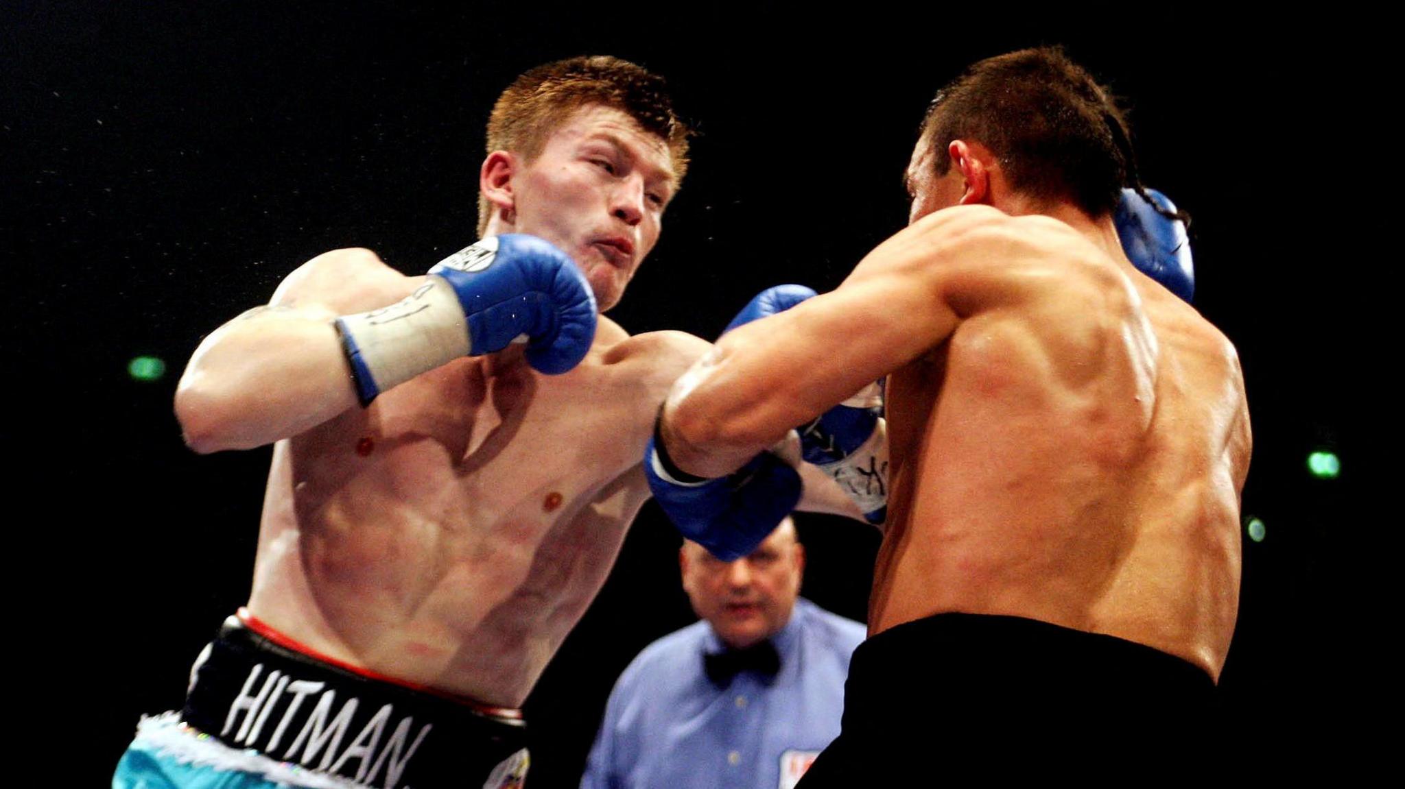 A mid shot of Ricky Hatton punching Kostya Tszyu. Both are wearing blue boxing gloves and shorts. In the background is the referee who is wearing a blue shirt and a dickie bow.
