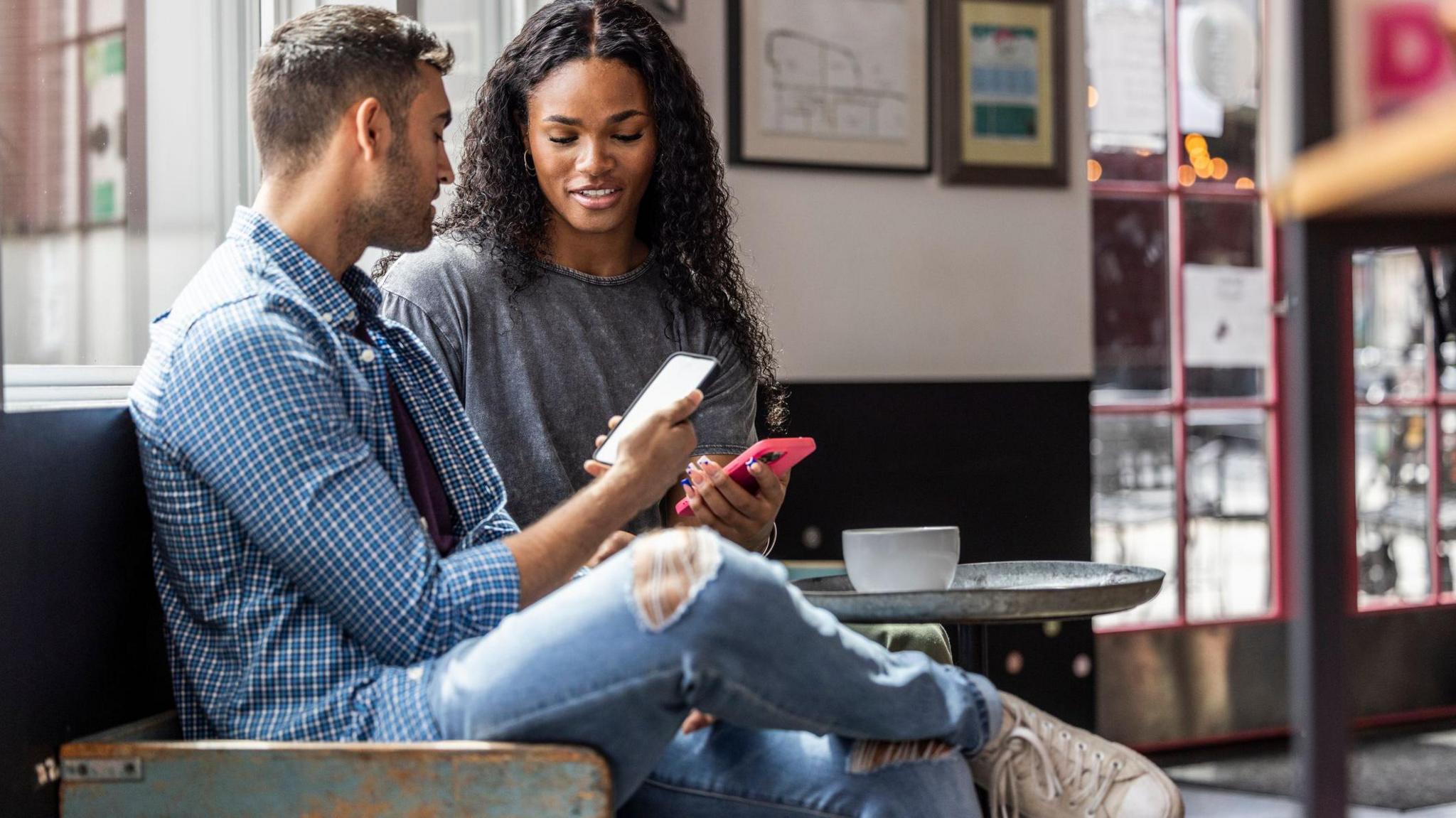 Man and a woman sit in a coffee shop each looking at a smartphone.
