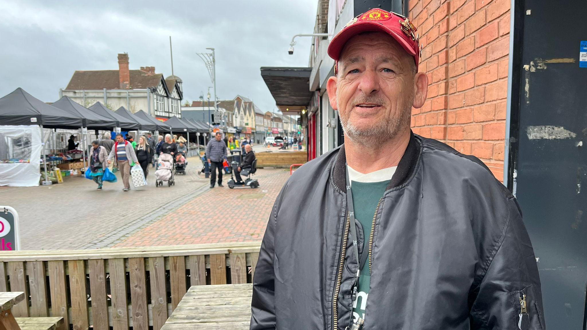 Philip Bastable in Bilston. He is wearing a black bomber jacket and a Manchester United hat. He has a vape around his neck. 