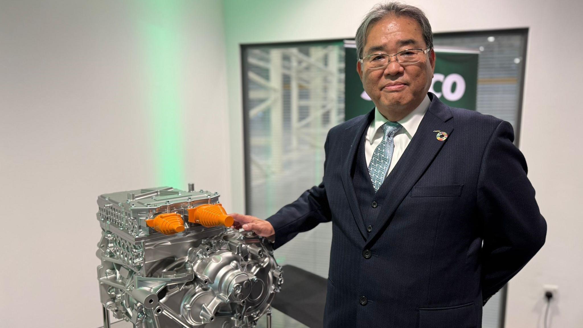 Tomoyoshi Sato standing alongside a powertrain. He has grey hair and is wearing glasses and a navy suit.