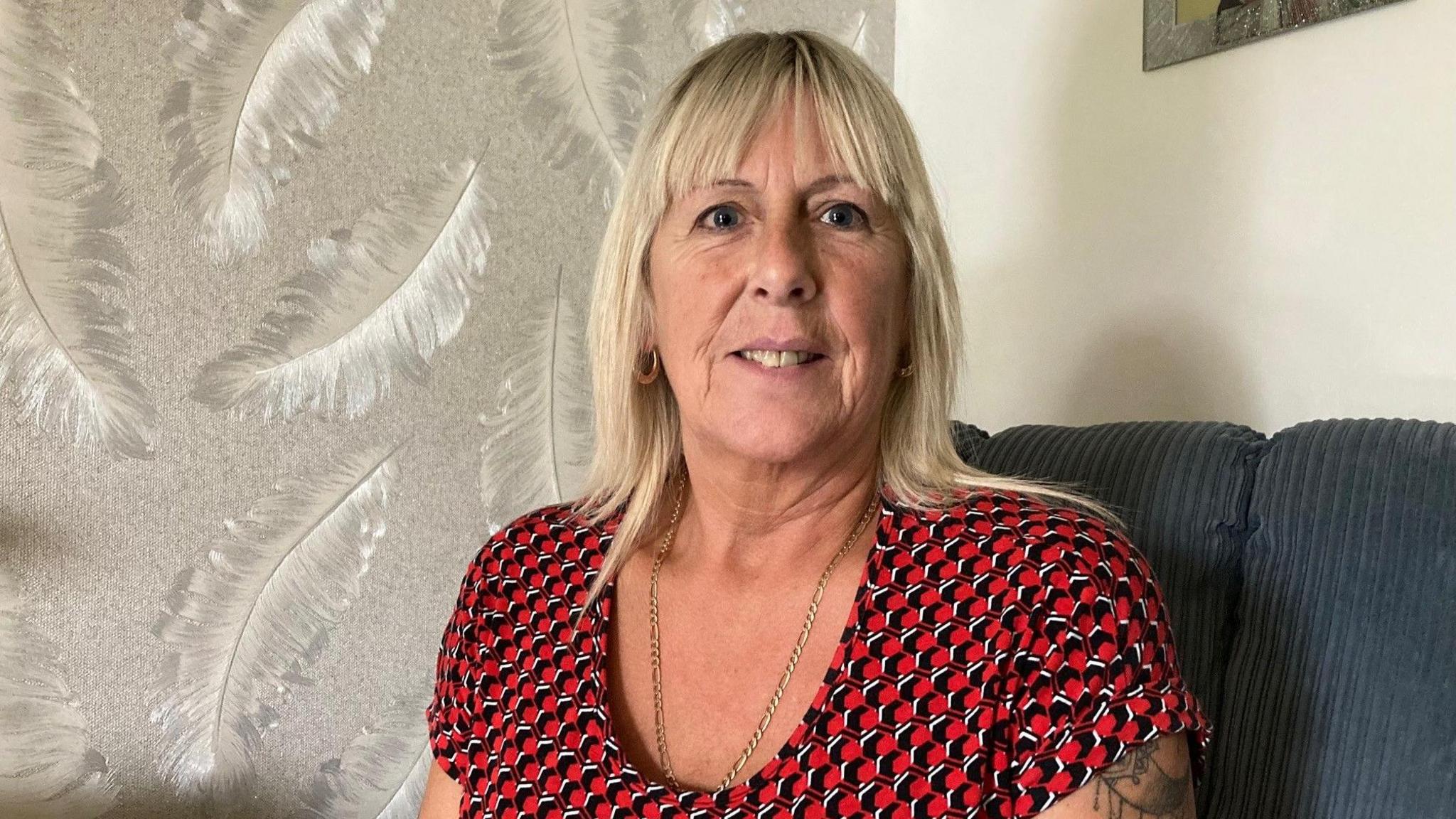 A blonde, middle-aged woman wearing a red, white and black sleeveless blouse with a tattoo showing on her left shoulder. 