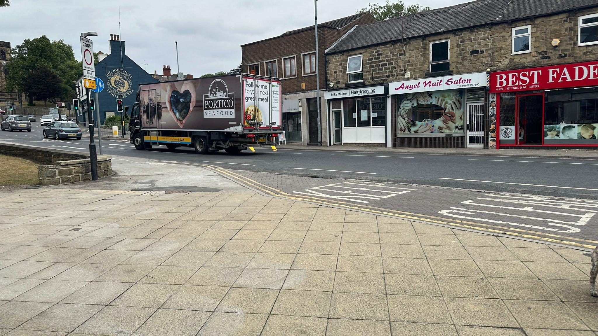 A paved pedestrian area alongside a road. There is a row of shops on the opposite side of the road. There is a lorry and cars travelling on the road.