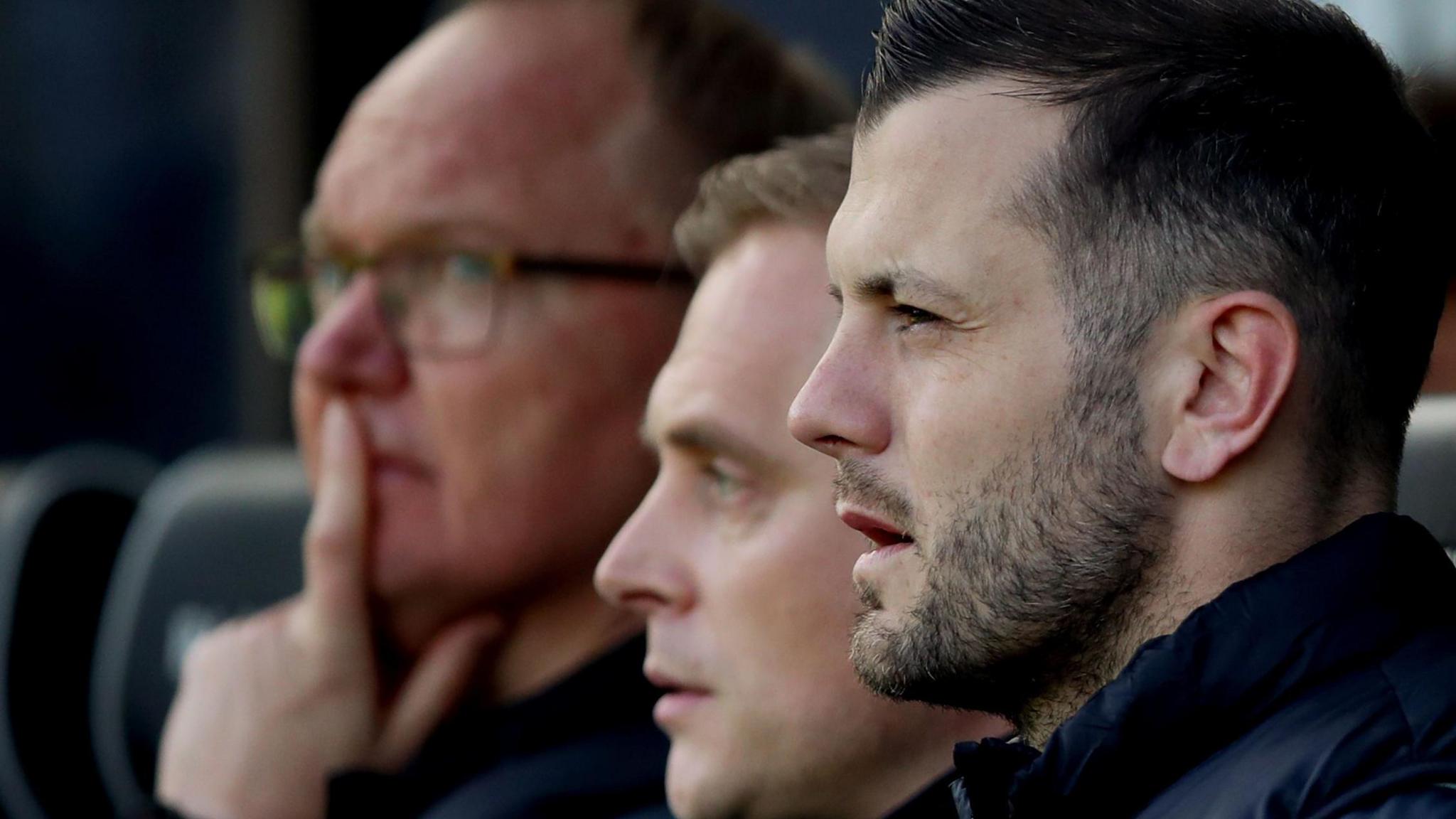 A close-up of Norwich City interim manager Jack Wilshere looking on during a match, with two men to his side. Wilshere has cropped dark hair and is wearing a puffa-style coat.