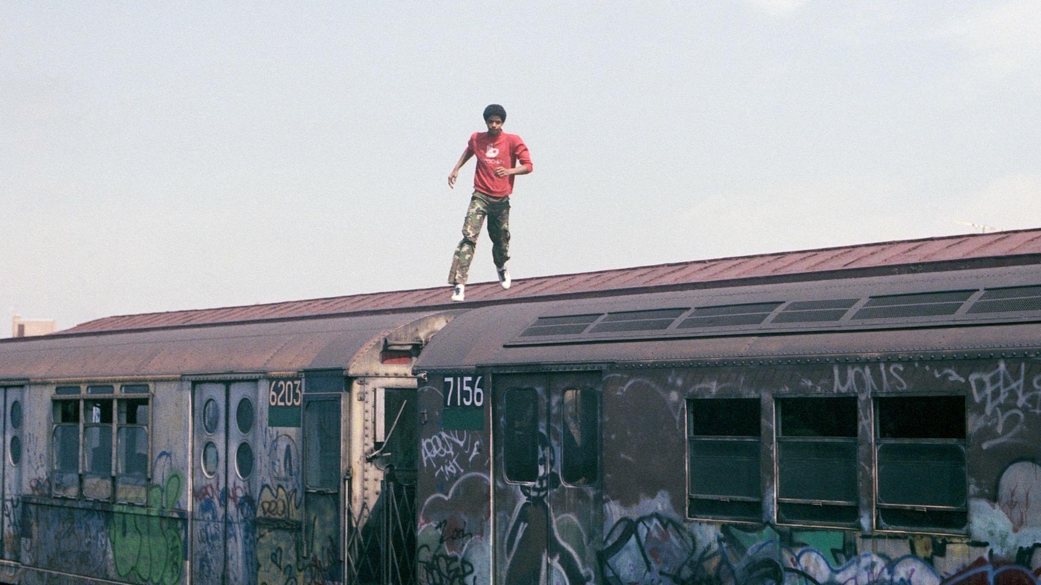 A man in a red sweatshirt and green trousers runs on top of a graffitied subway train carriage on tracks.
