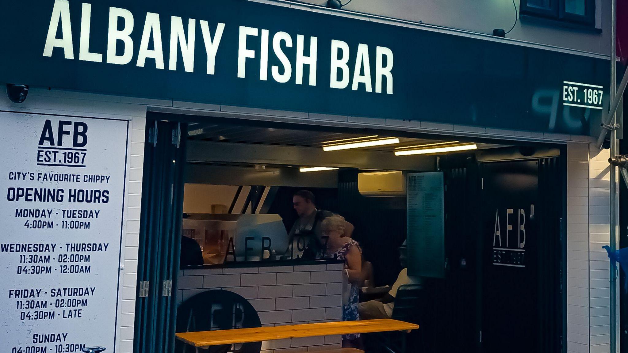 Albany Fish Bar sign, established 1967, above an open takeway with a man a woman at the counter