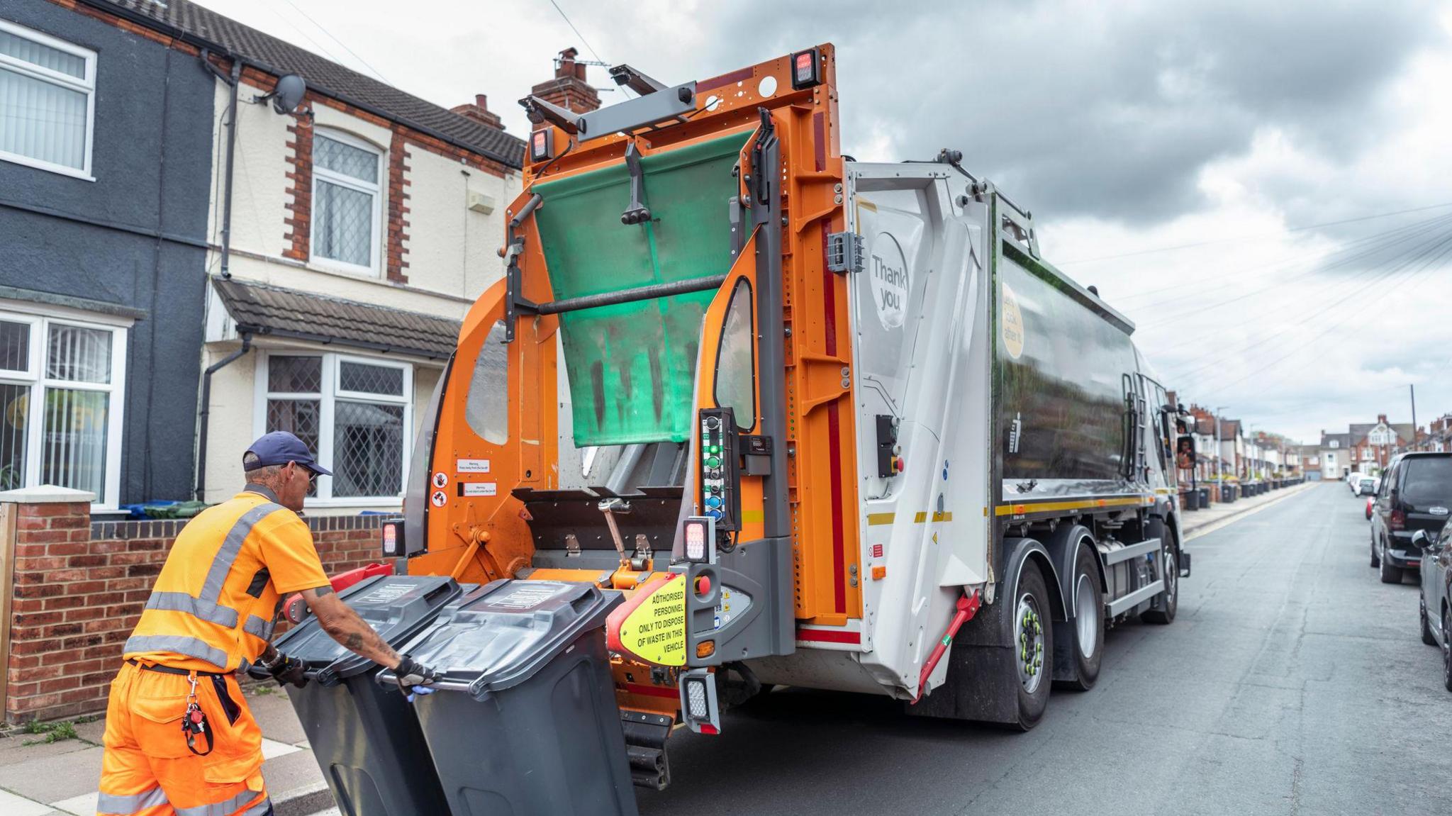 A refuse collector pushing two bins towards a bin lorry in a suburban street. Various houses, including a grey-fronted building on the left and a number of parked cars can also be seen.