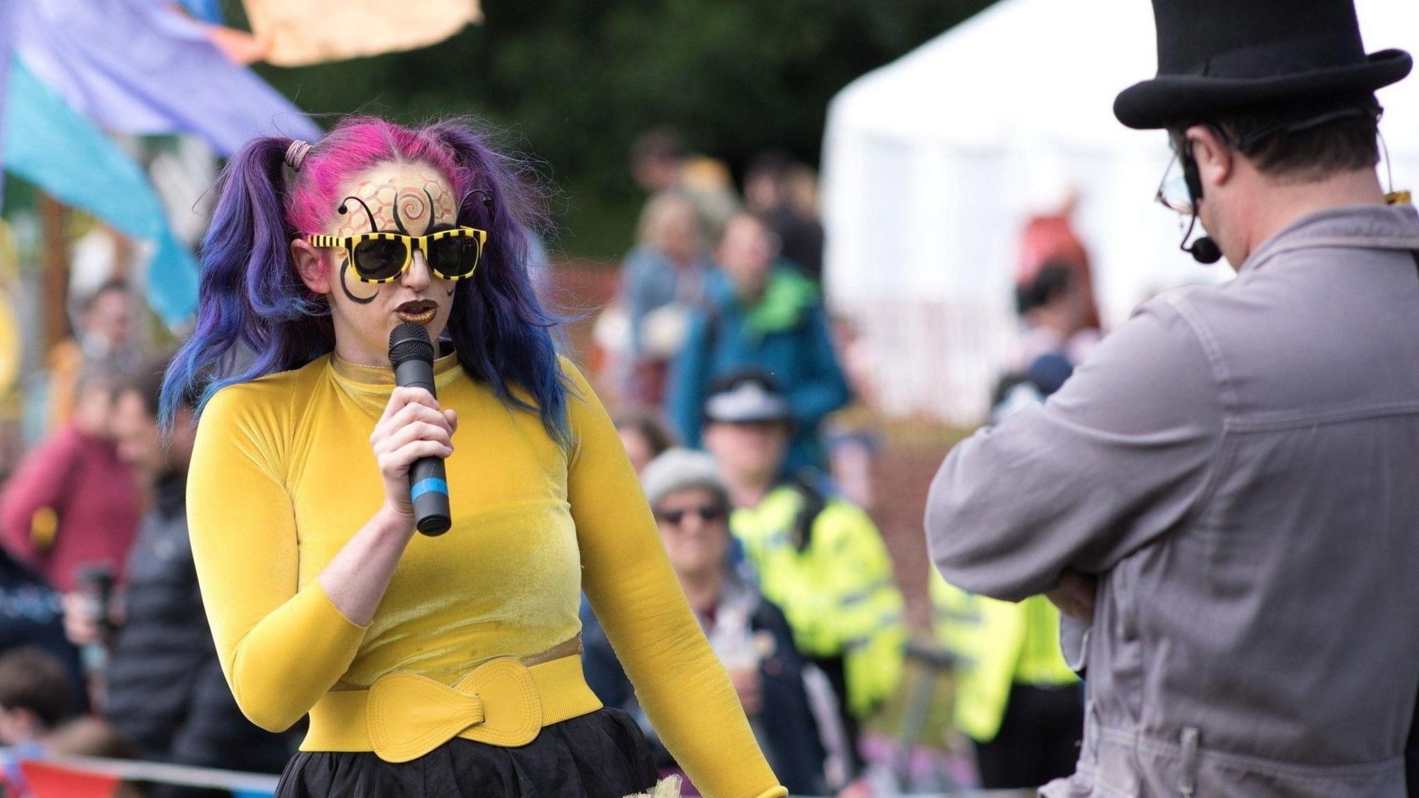 A woman with two hair bunches that are dyed pink and purple is singing into a microphone. She is wearing black and yellow stripy sunglasses and has black and orange honeycomb style face paint. She is wearing a yellow top and belt