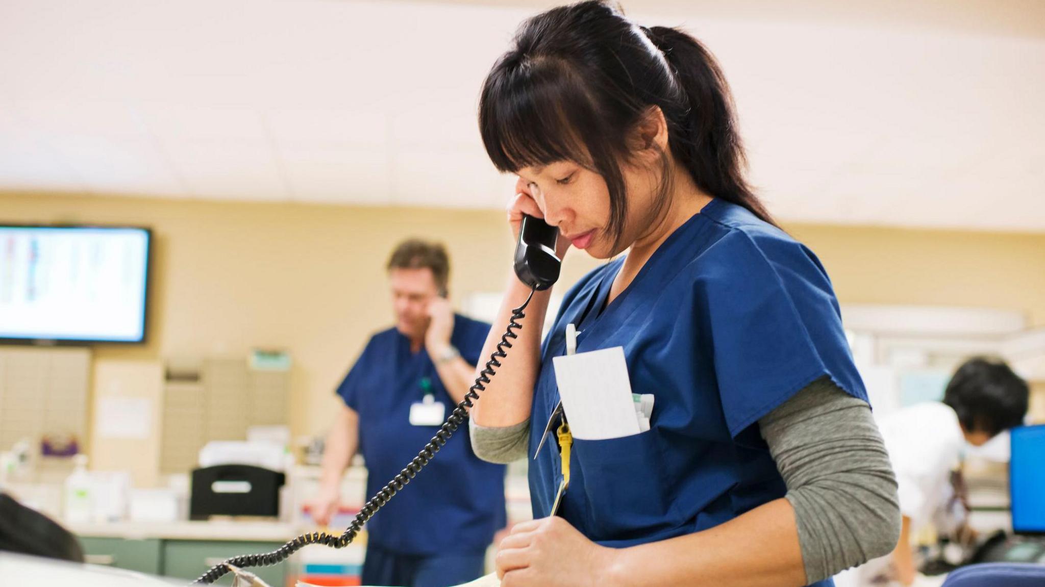 a woman in nursing scrubs on the phone in a reception setting, with other uniformed staff in the background