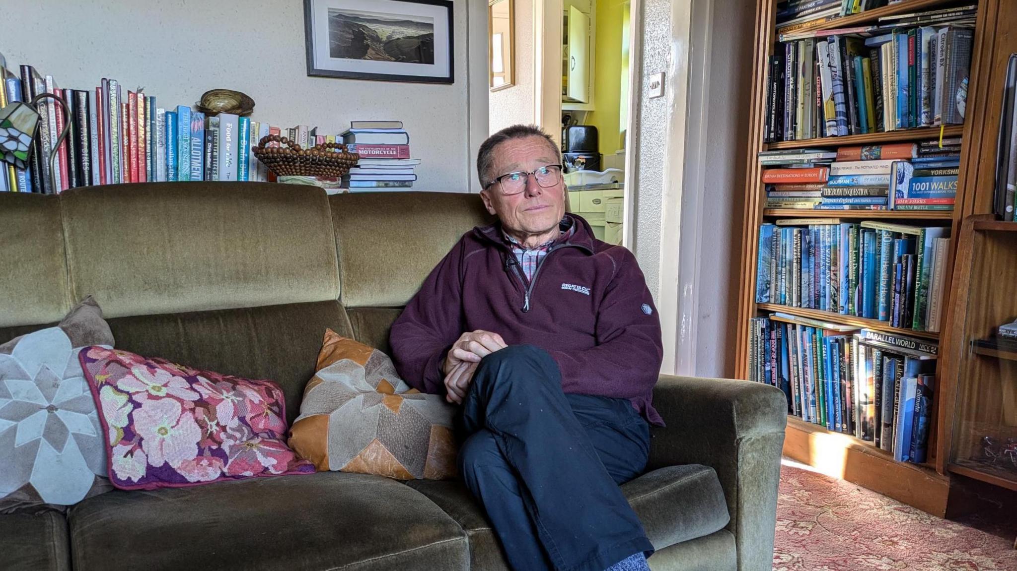 Andrew, a man with short grey hair and glasses stares away from the camera and into the middle distance. He is wearing a burgundy zipped jumper and sitting on a brown sofa in his living room.